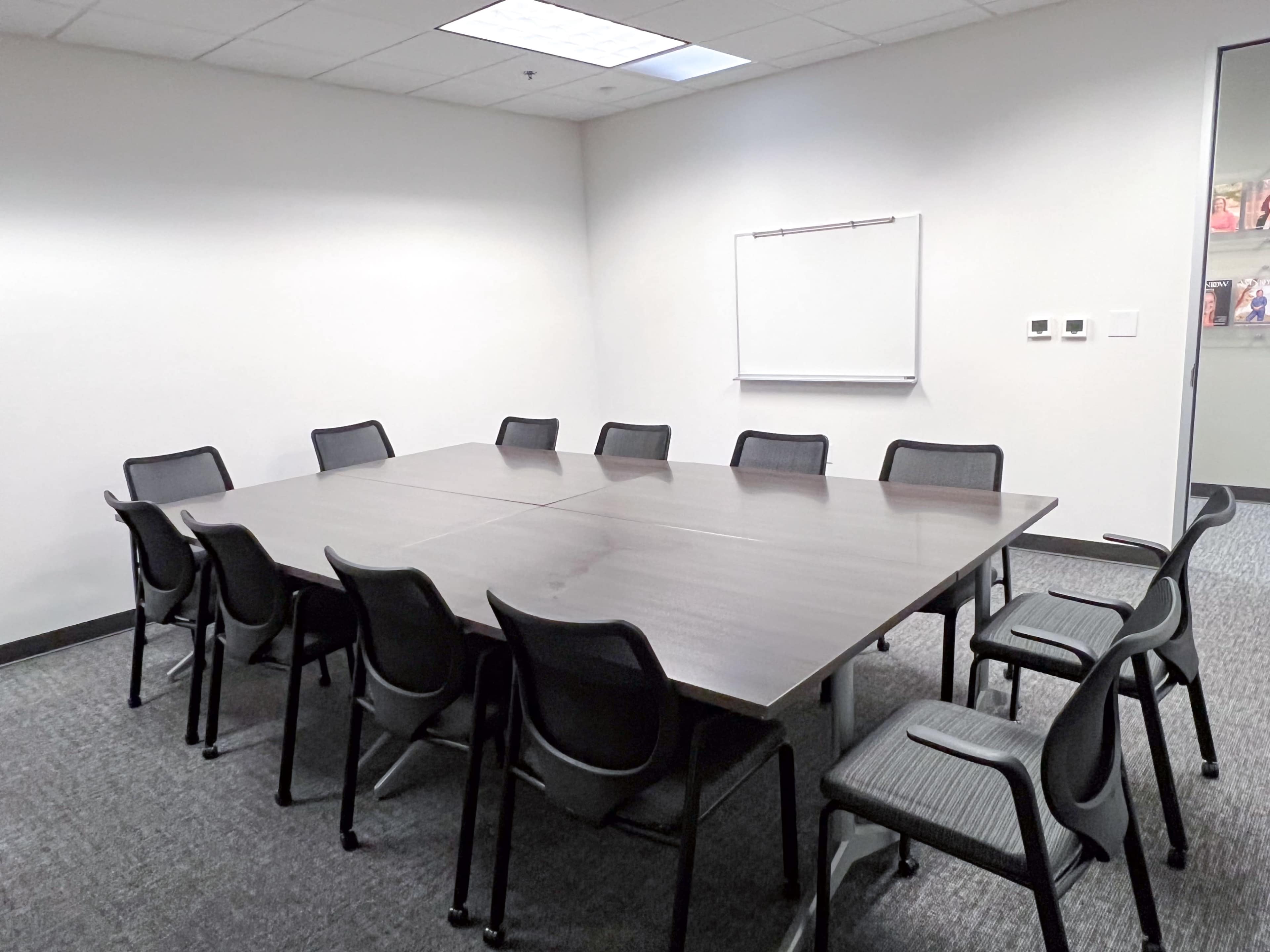 A large conference room features a rectangular table surrounded by twelve chairs, with a whiteboard mounted on the wall.