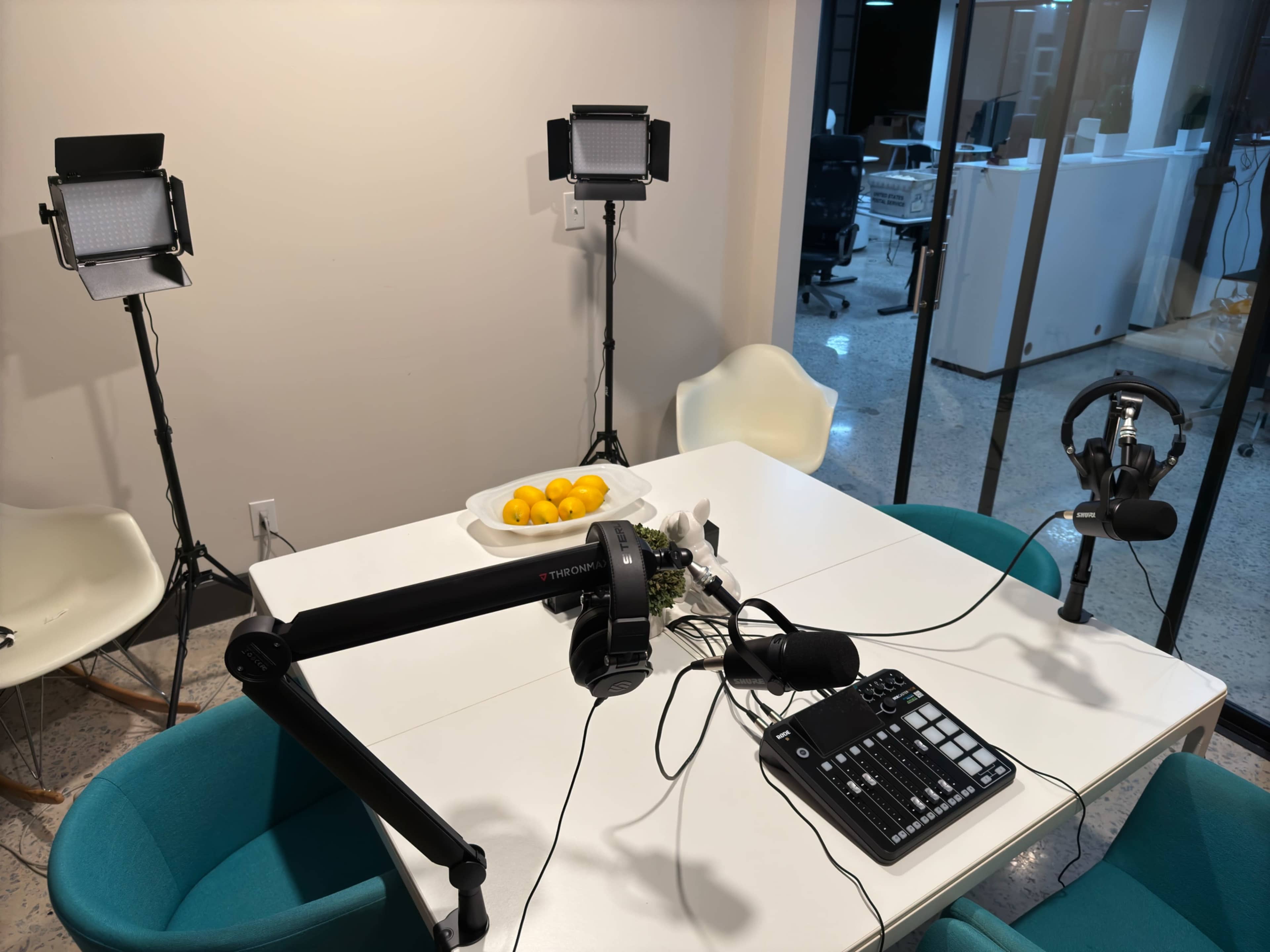 A podcasting setup is arranged on a white table featuring microphones, headphones, and studio lights, with a bowl of lemons in the center.