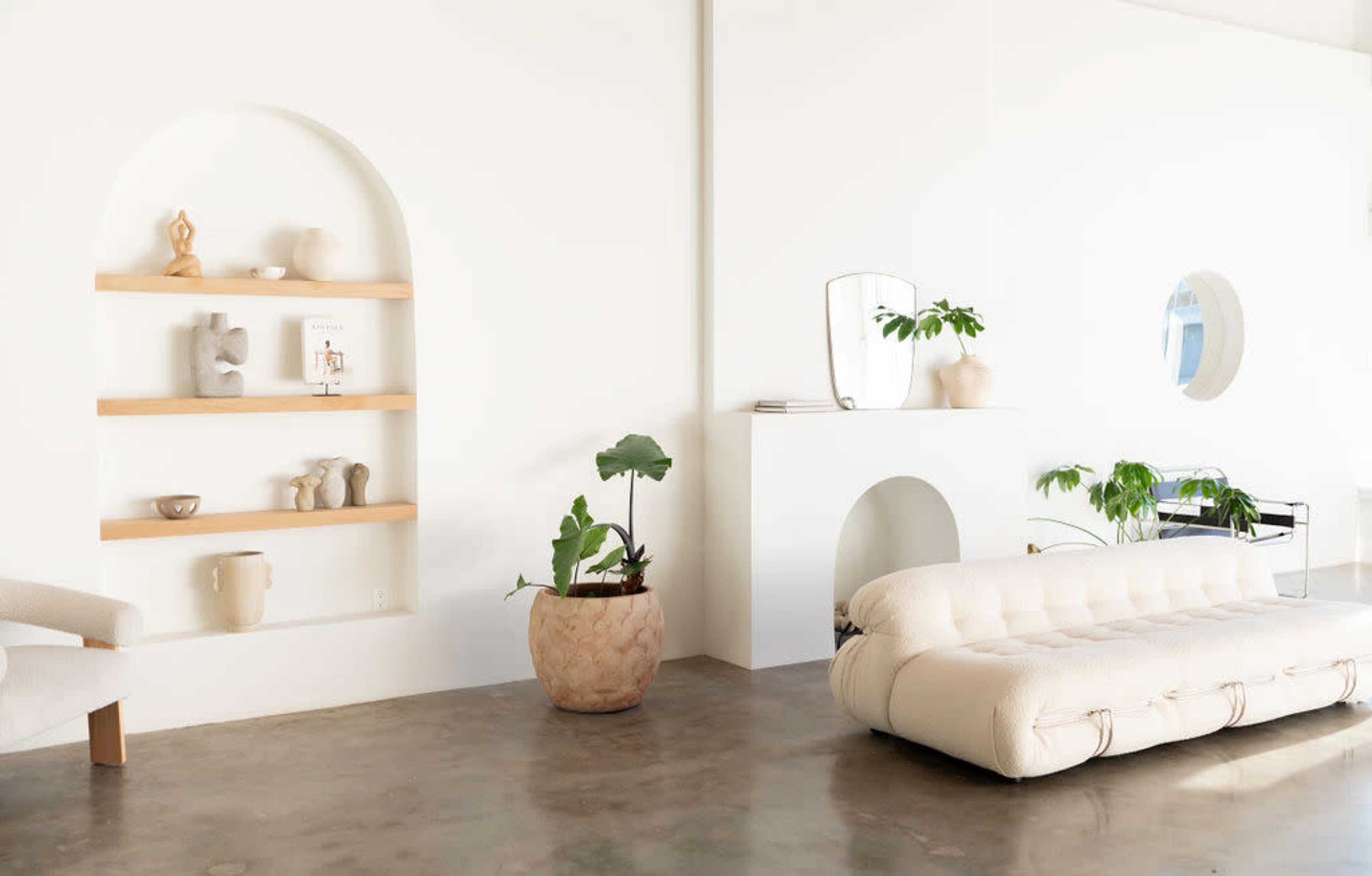 The image shows a minimalist interior with a white wall, a shelf displaying various decorative items, and a light-colored sofa on a polished concrete floor.
