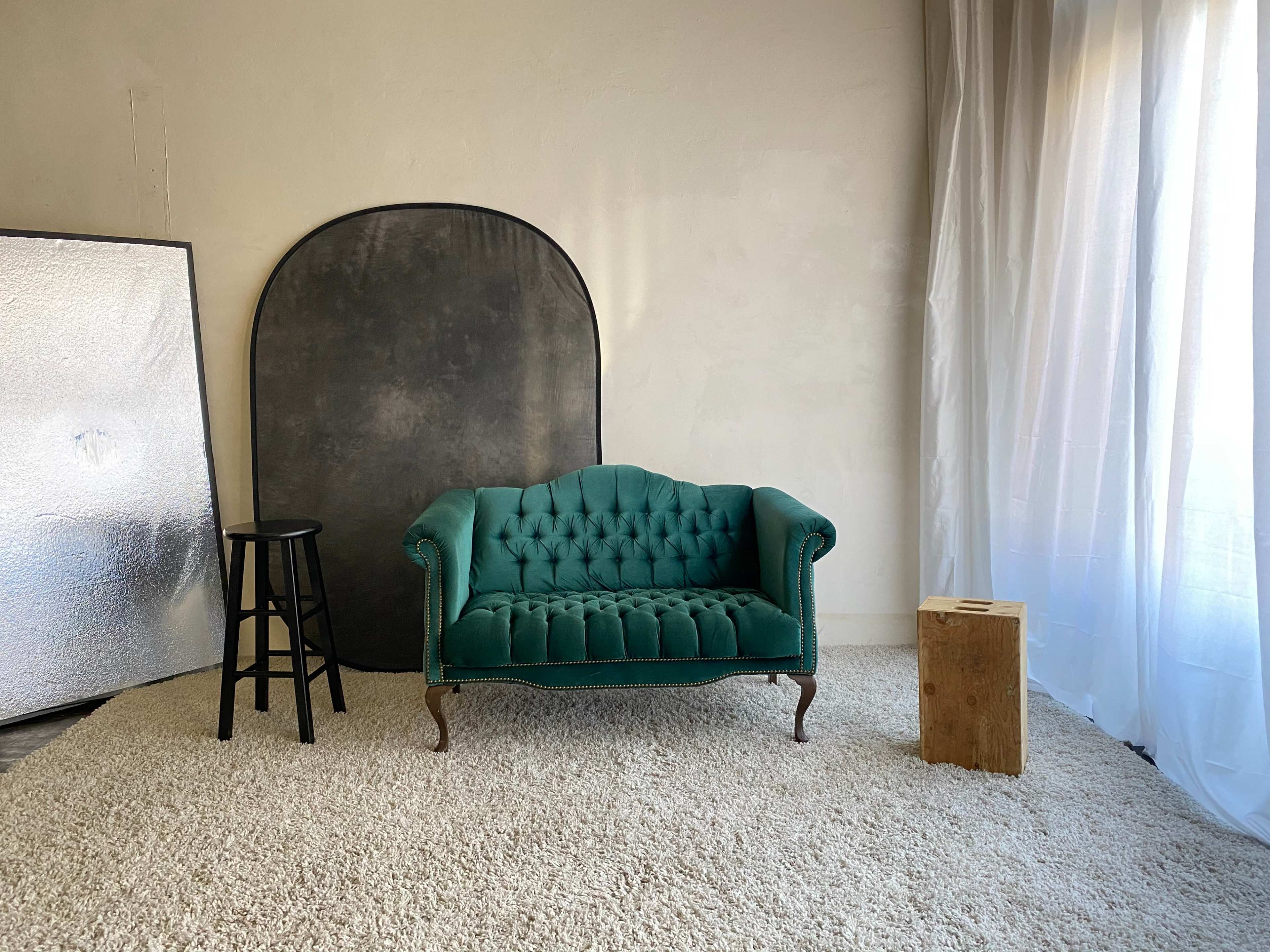 A green tufted couch, a black stool, a wooden block, and a backdrop in a room with white curtains and a light-colored wall.