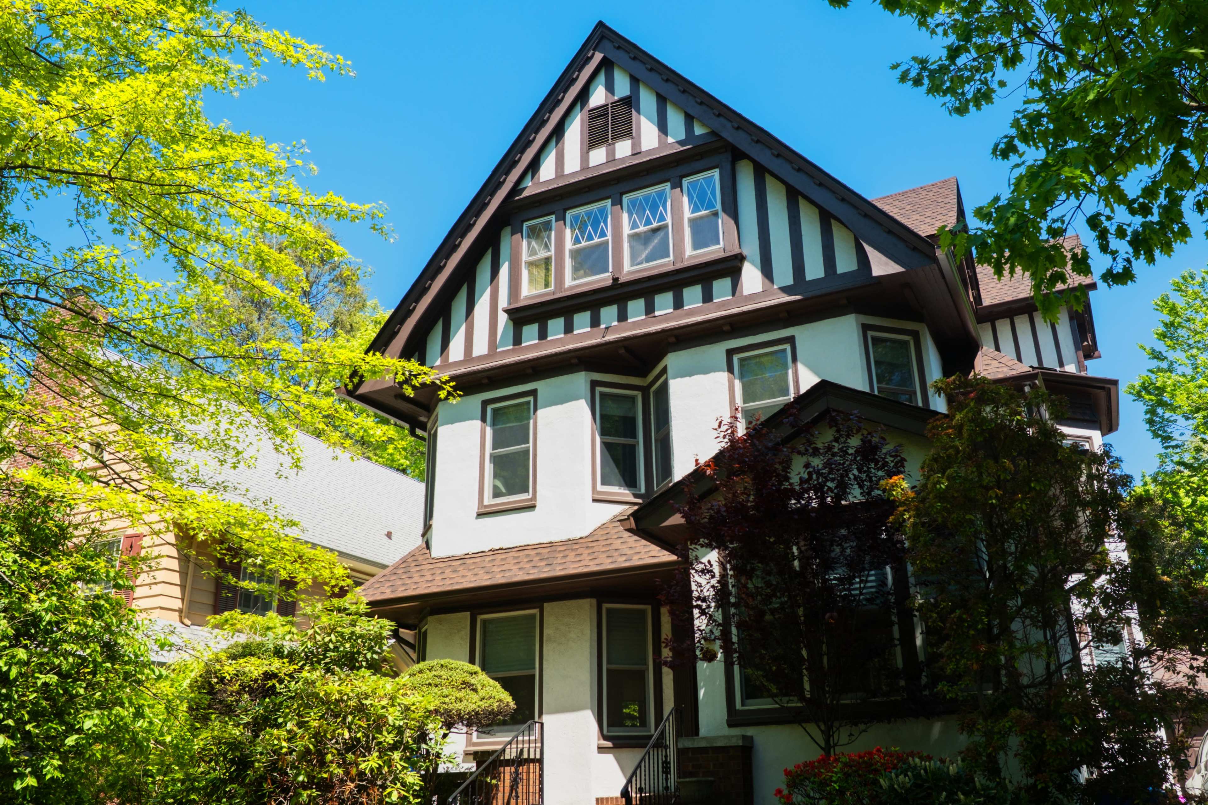 The image shows a two-story house with a steep gabled roof and distinctive half-timbering on the upper level, surrounded by lush greenery.