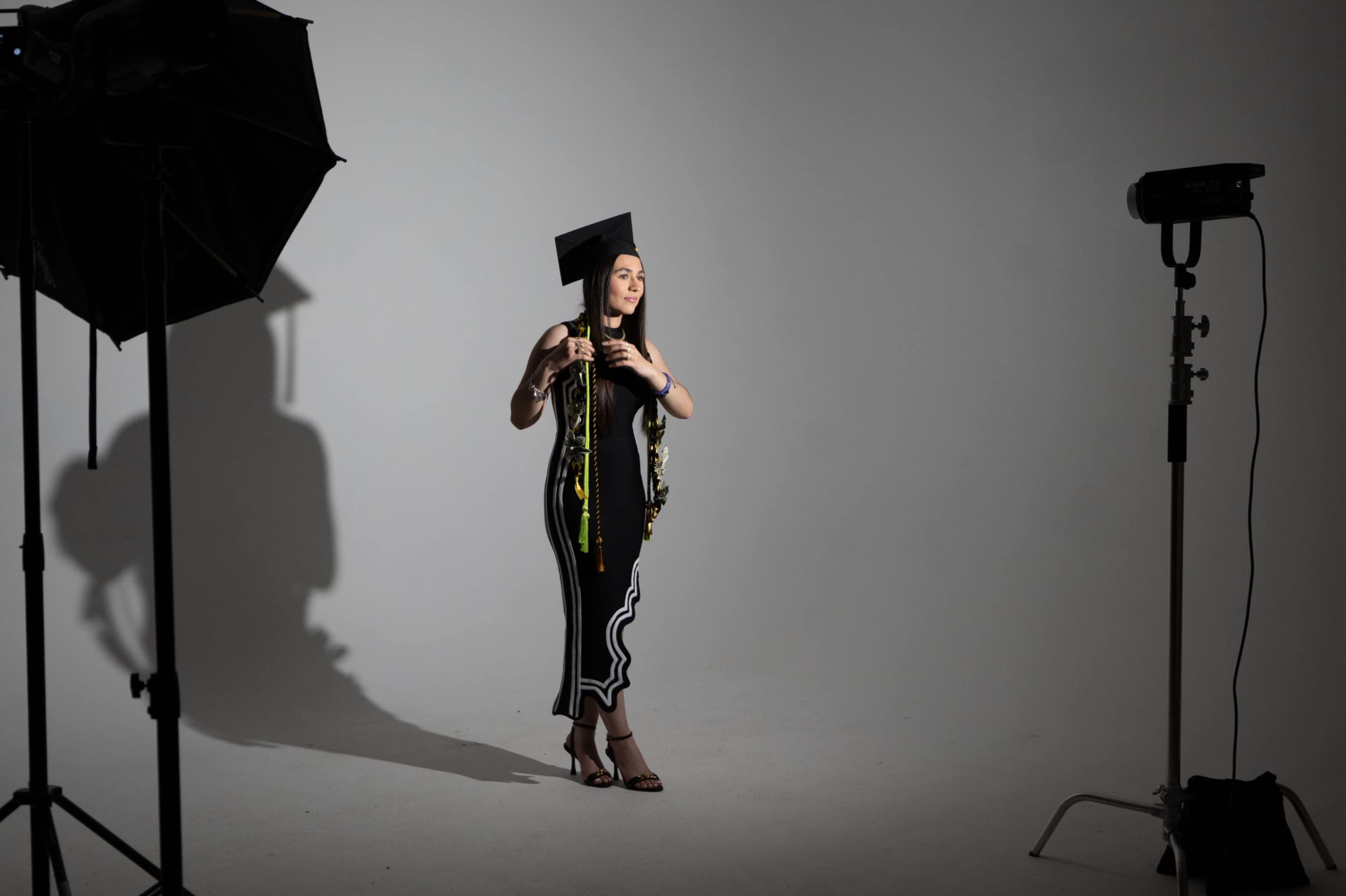 A graduate in a black cap and gown stands in a studio, holding a tassel and wearing a lei, with lighting equipment positioned nearby.