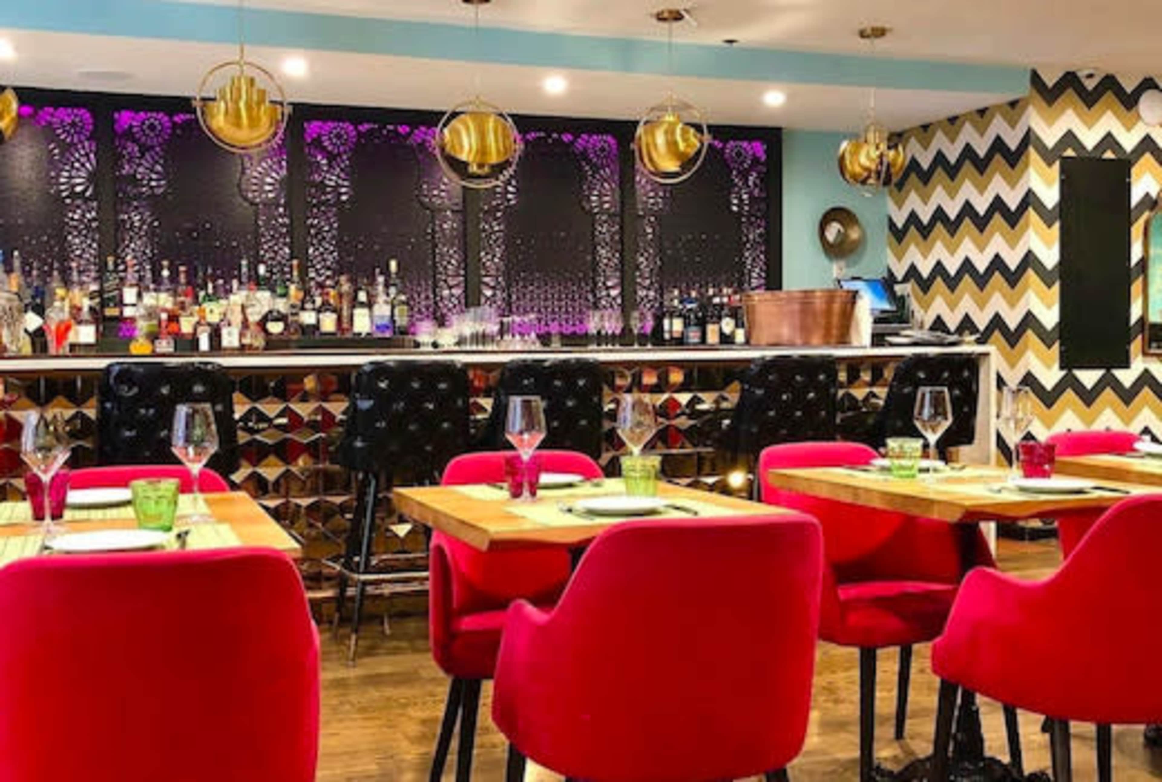 The image shows a modern restaurant interior with red upholstered chairs, a wooden dining table set for guests, and a bar area featuring a variety of liquor bottles in the background.