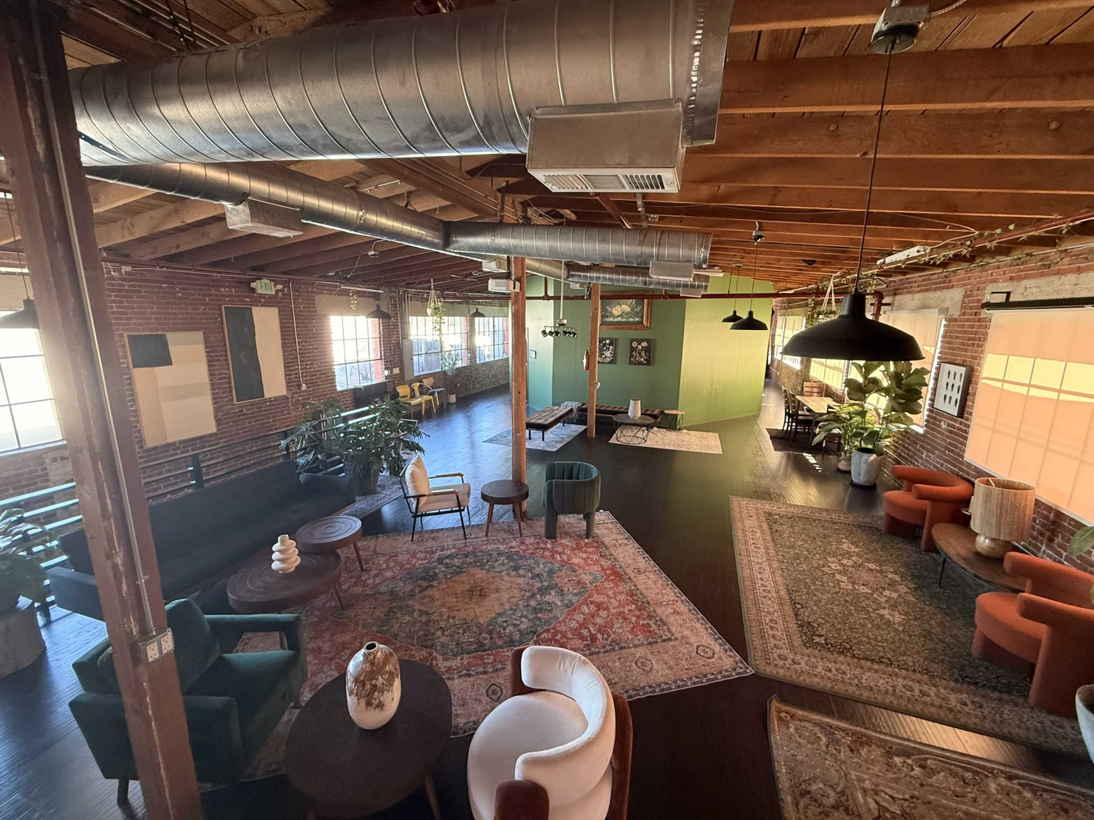 The image shows a spacious, industrial-style interior with exposed wooden beams, large windows, various seating areas, and decorative rugs.