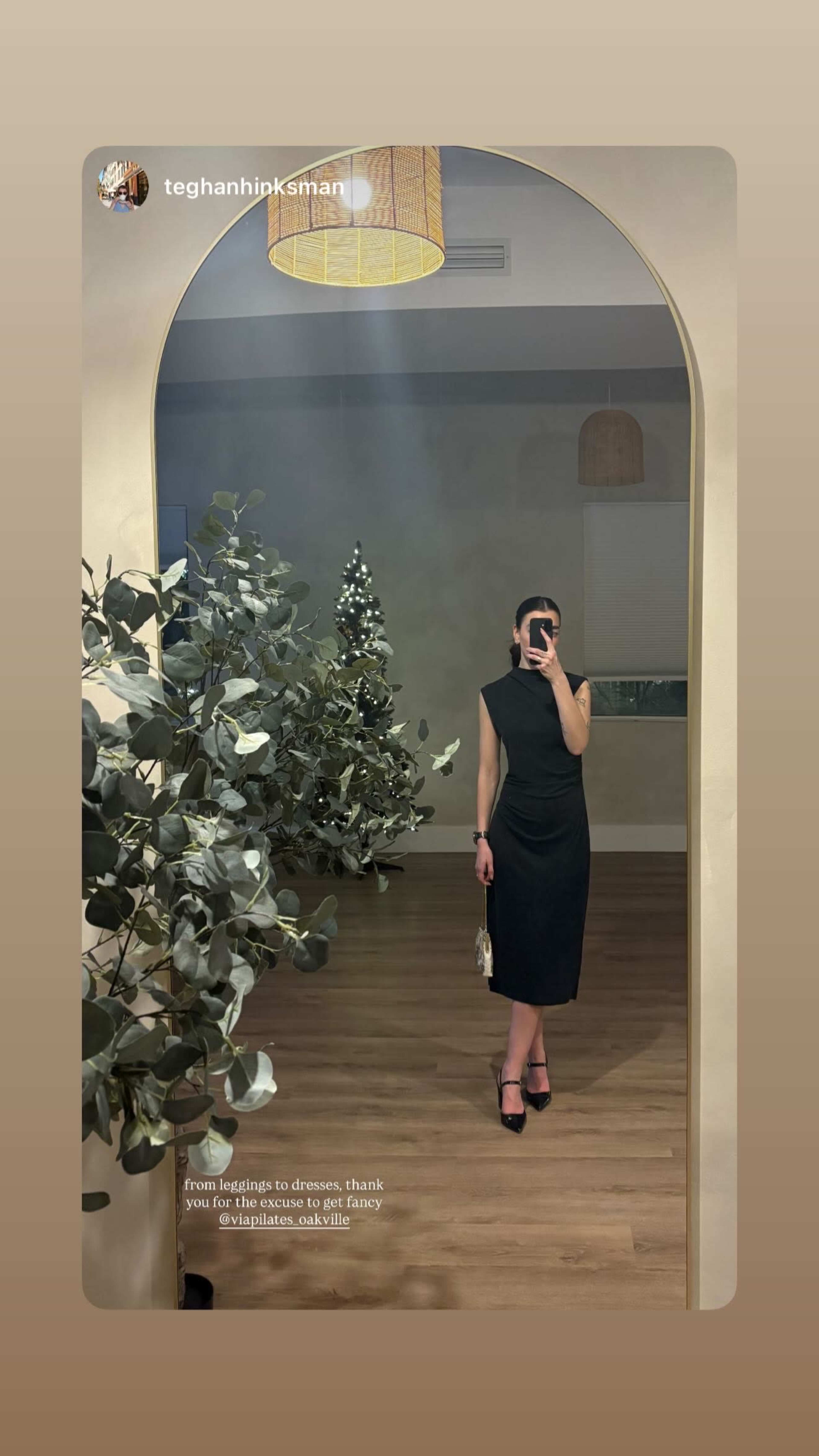 A person in a black dress stands in front of a mirror, surrounded by decorative plants and a Christmas tree in a well-lit room.