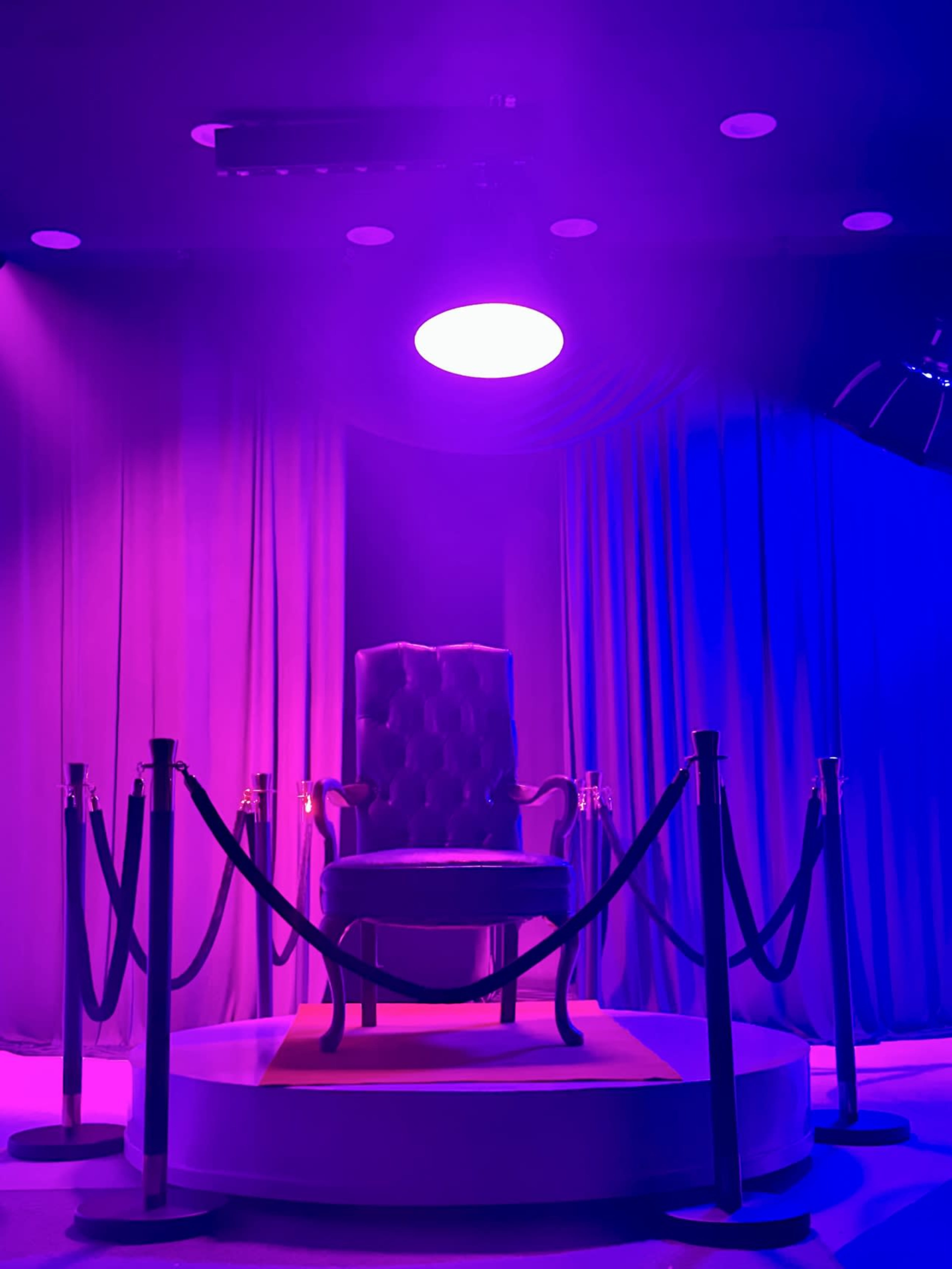 The image shows a spotlight above a single velvet chair surrounded by stanchions and purple drapery.
