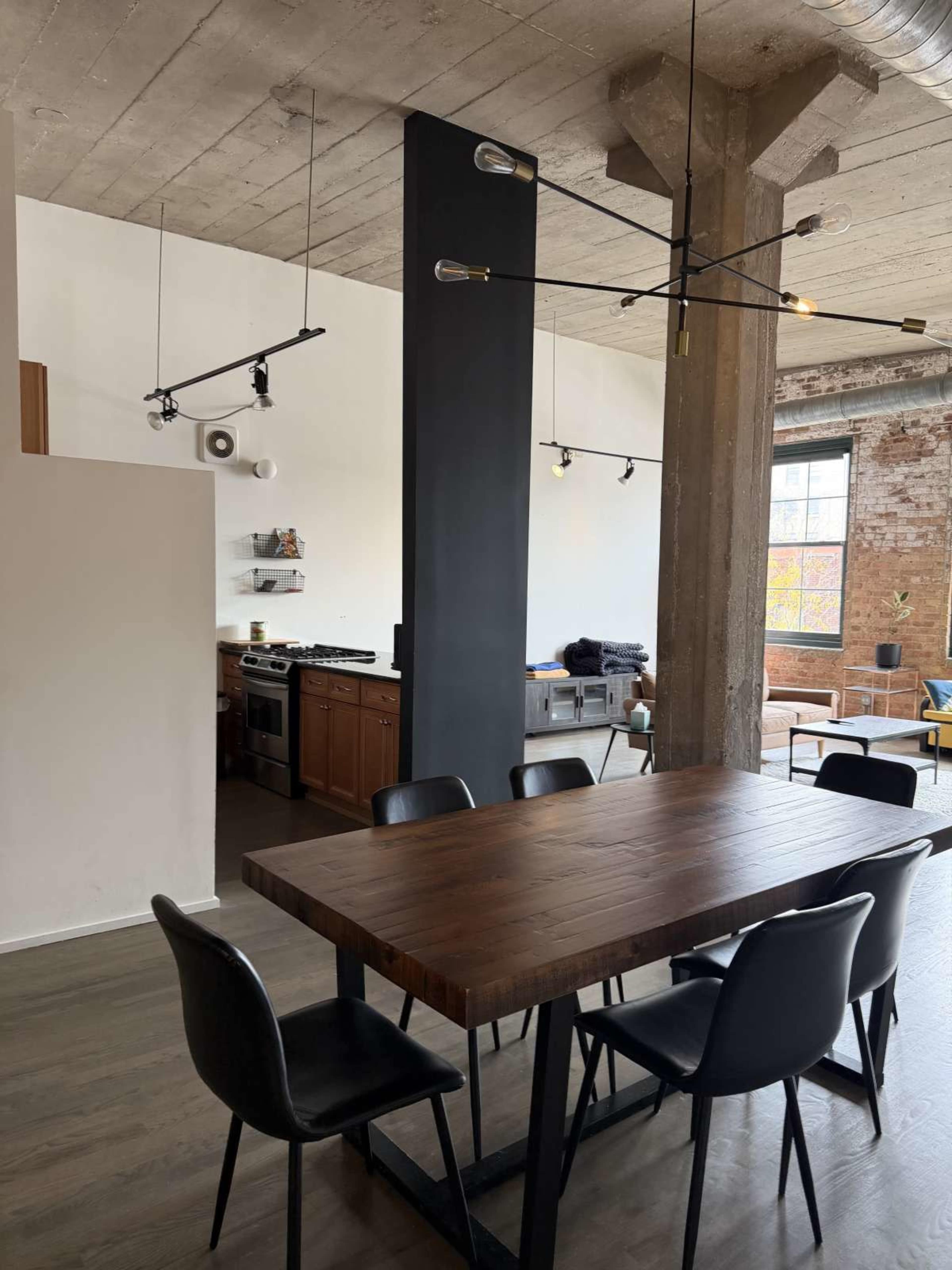 The image shows a modern dining area with a wooden table and black chairs, adjacent to a kitchen space featuring wooden cabinetry and industrial-style lighting.