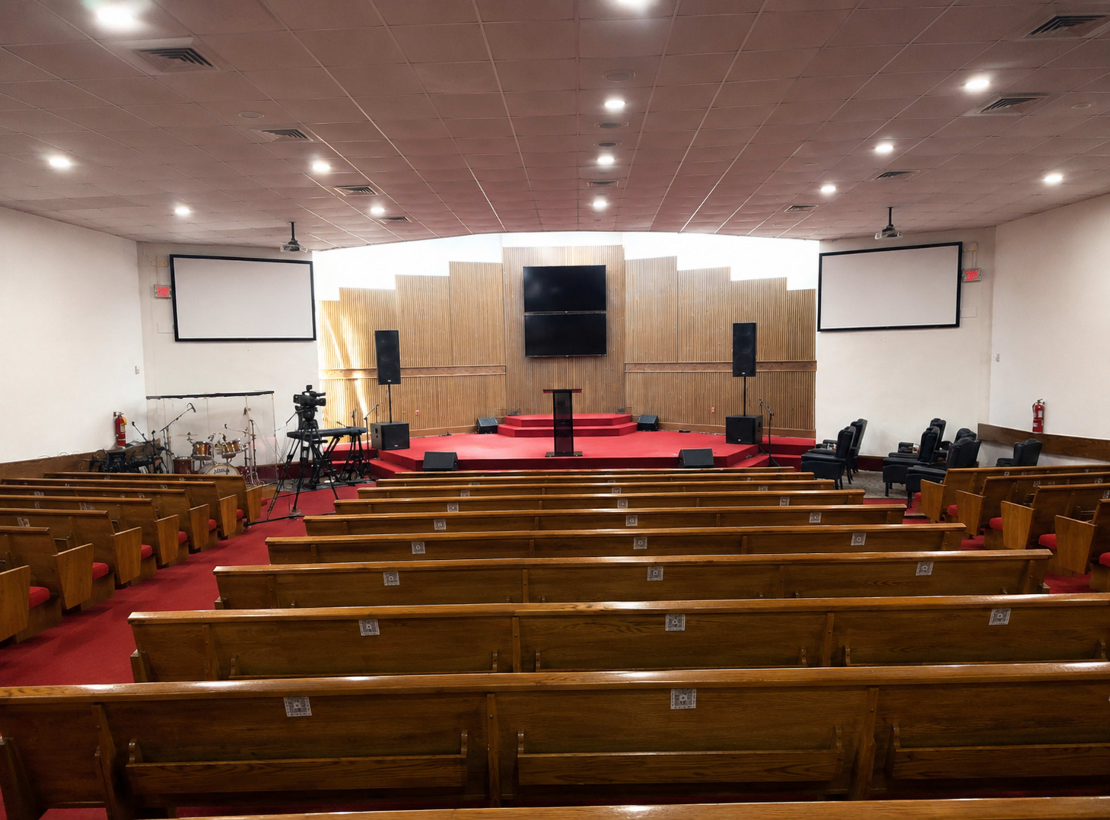 The image shows the interior of a church, featuring wooden pews arranged in rows facing a stage with a podium and audio-visual equipment.