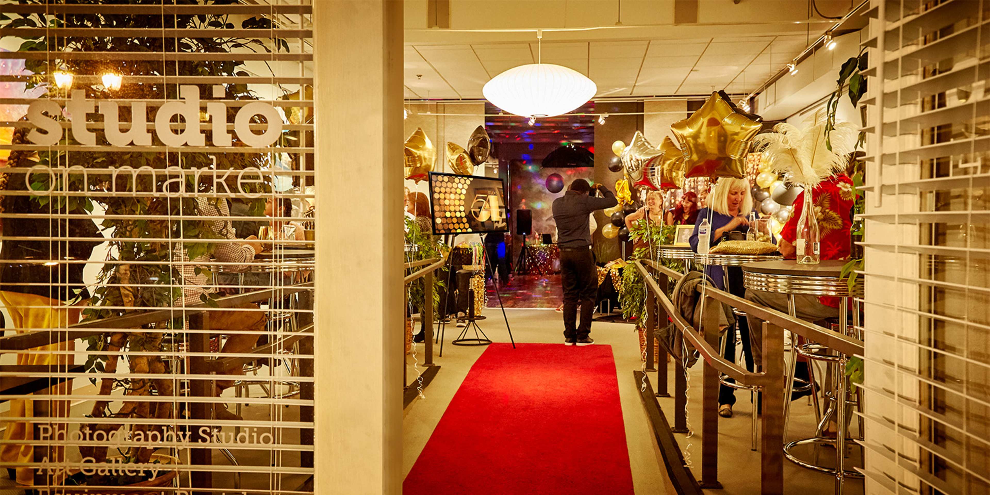 The entrance of a photography studio is decorated with balloons and gold stars, leading to a red carpet and a gathering of guests inside.