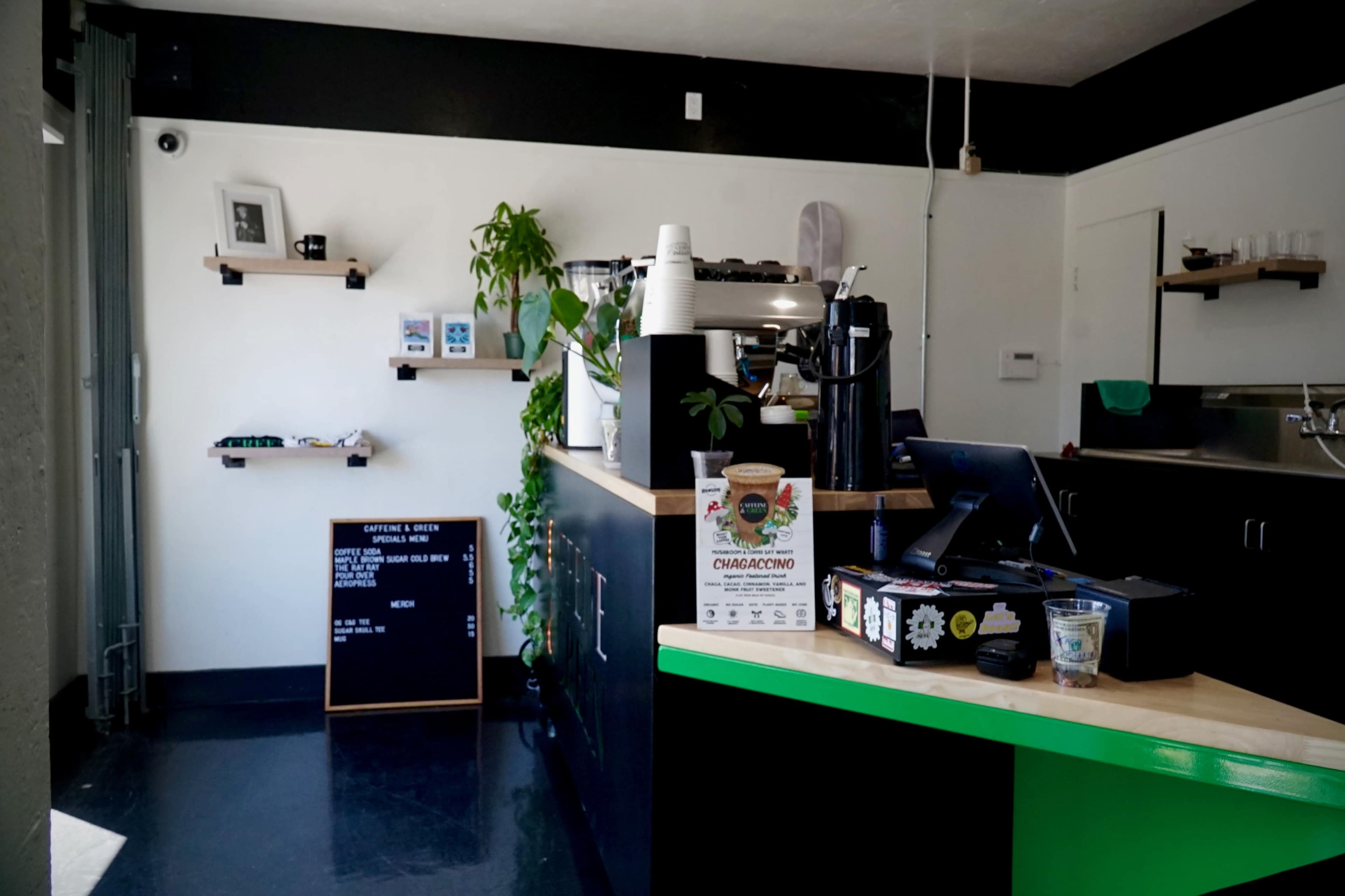 A coffee shop interior features a minimalist counter with a register, coffee machine, and plants, alongside a chalkboard menu and shelves displaying items.