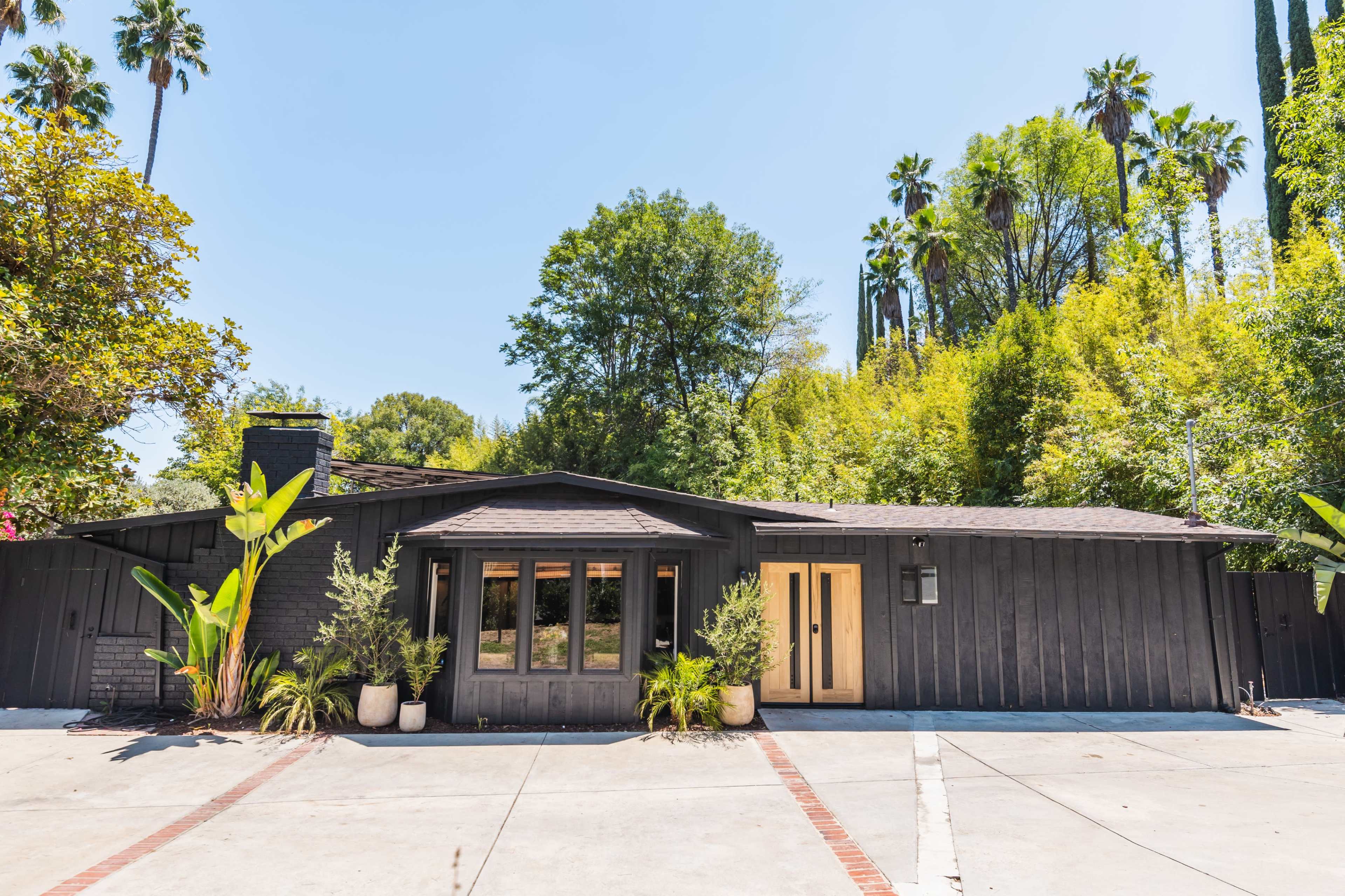 A black wooden house with large front windows is surrounded by lush greenery and palm trees.