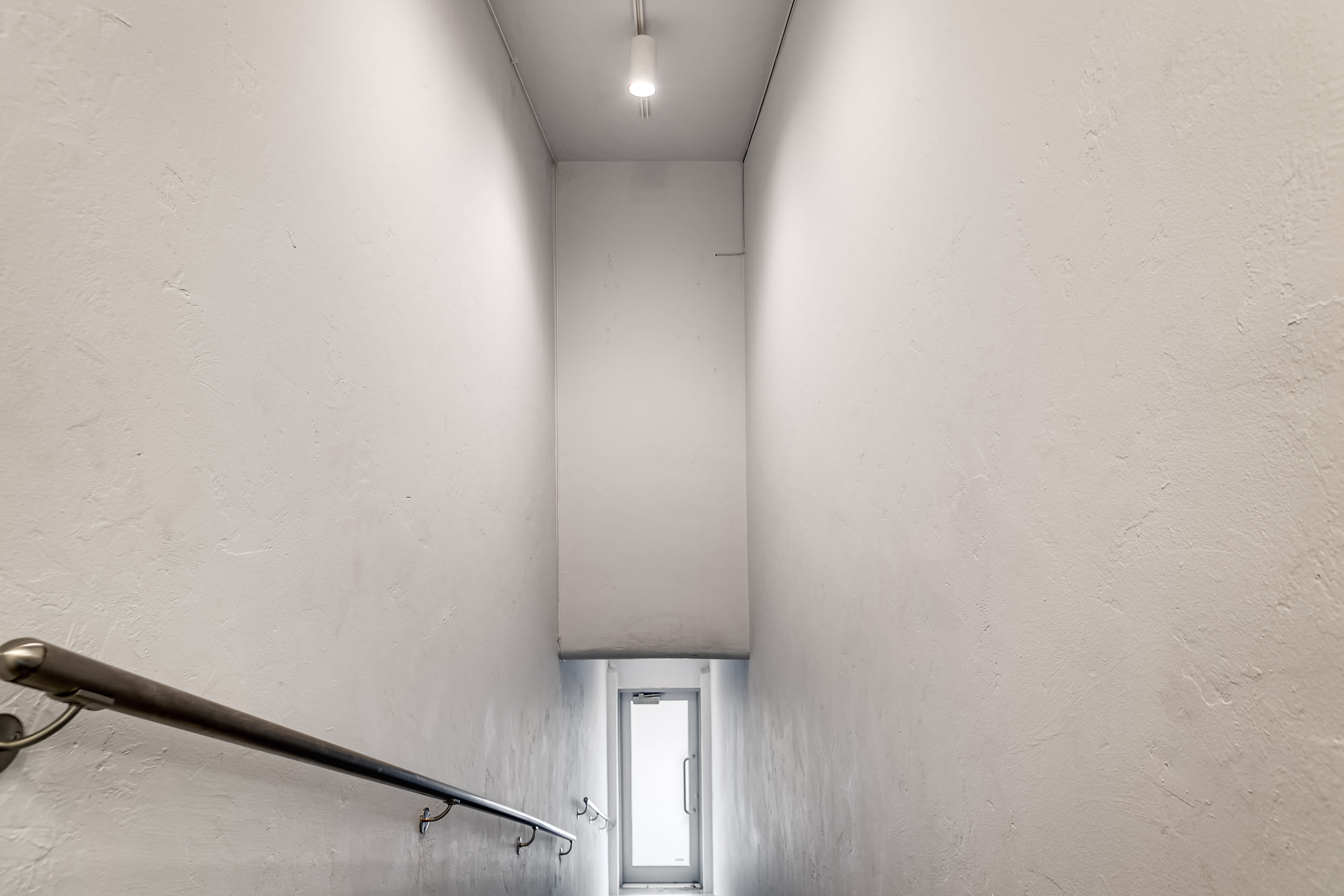 A narrow, well-lit staircase leads down to a white-walled landing with a door at the bottom.