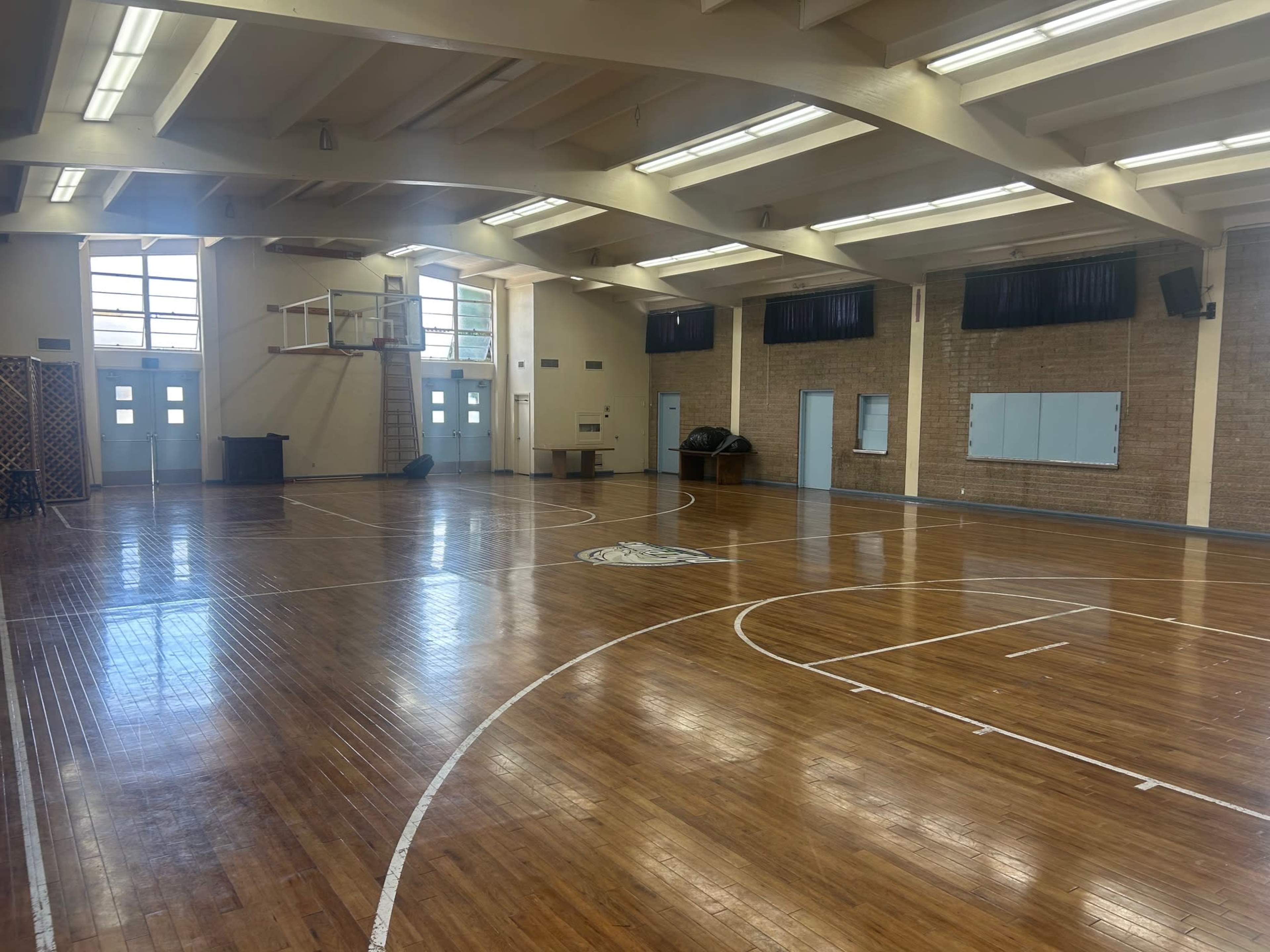 The image shows an empty gymnasium with polished wooden floors, large windows, and basketball hoops mounted on the walls.