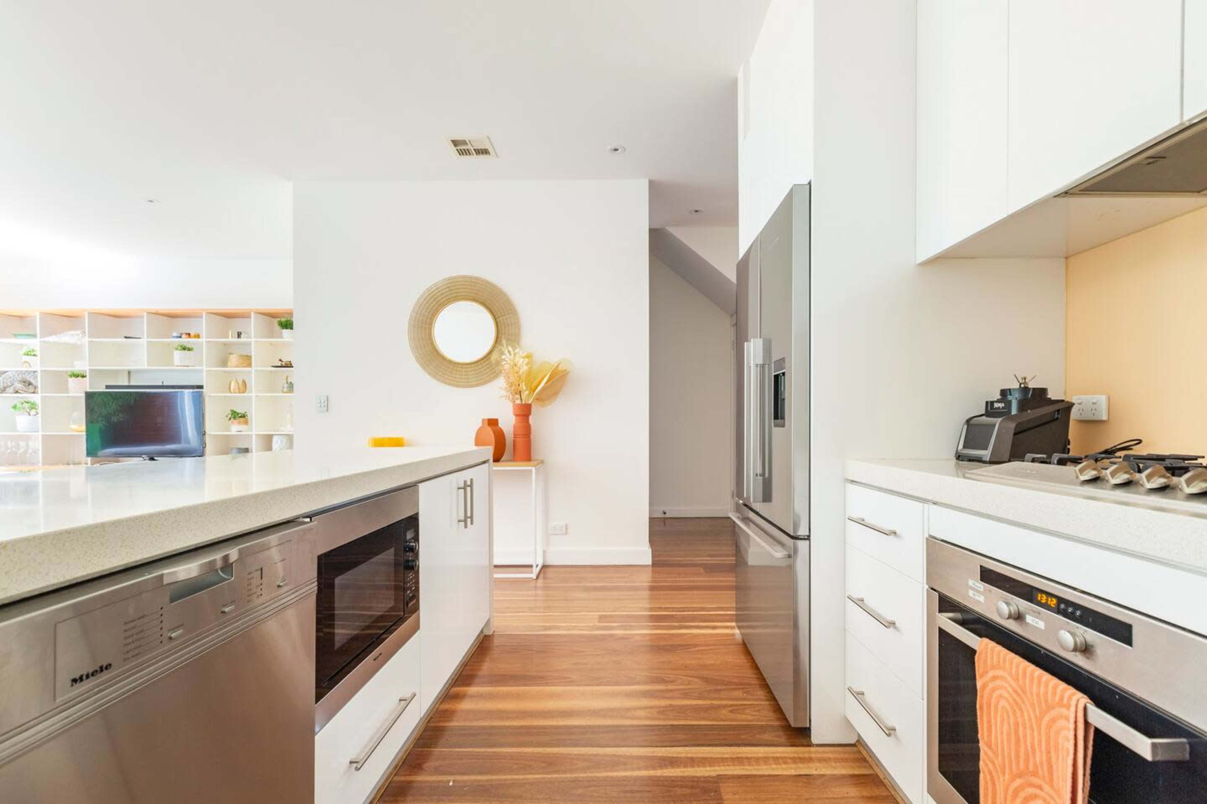 A modern kitchen features a central island with a microwave and oven, stainless steel refrigerator, and wooden flooring, leading to an open living area.