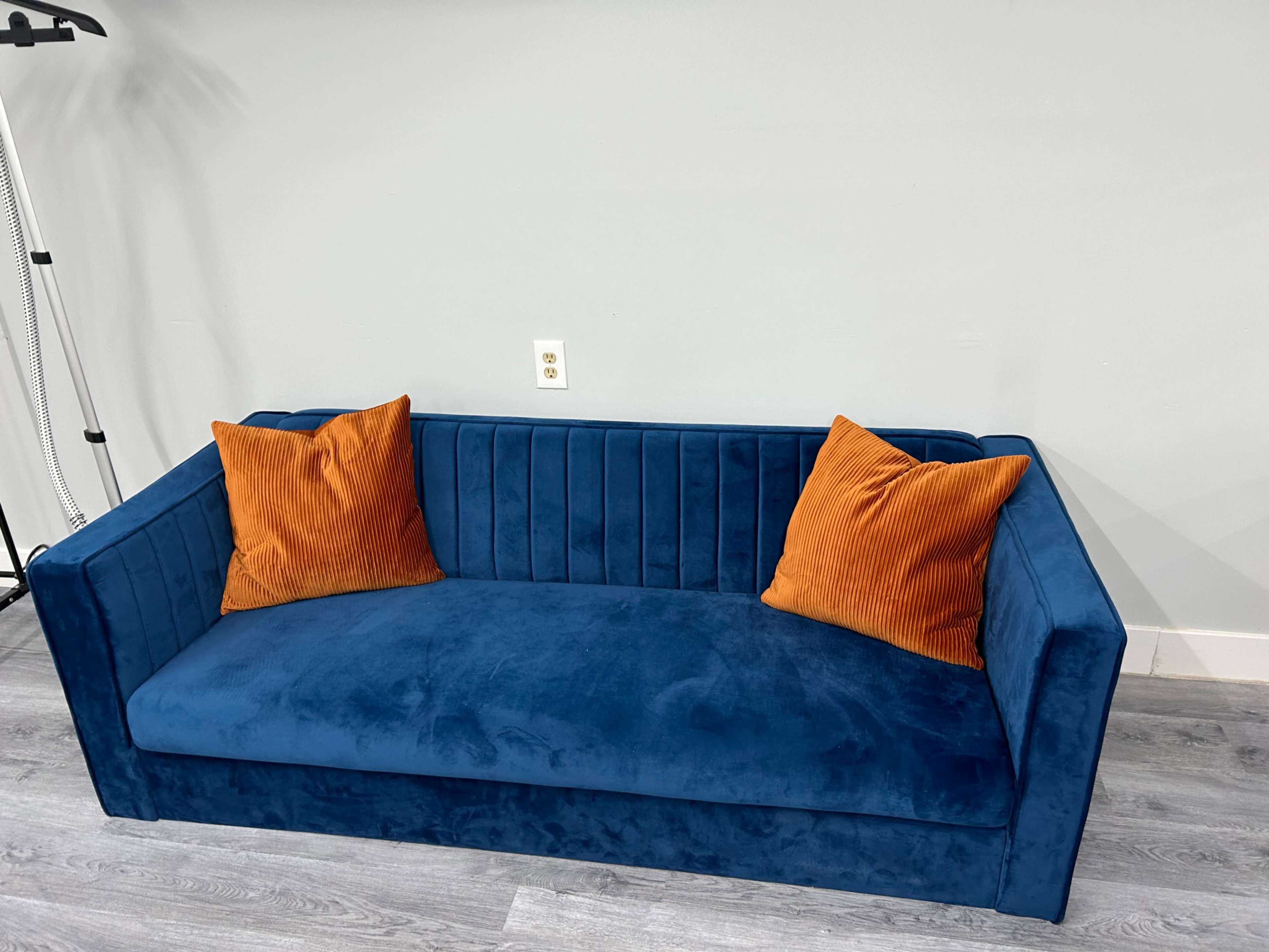 A blue velvet sofa with orange striped cushions is positioned against a gray wall.
