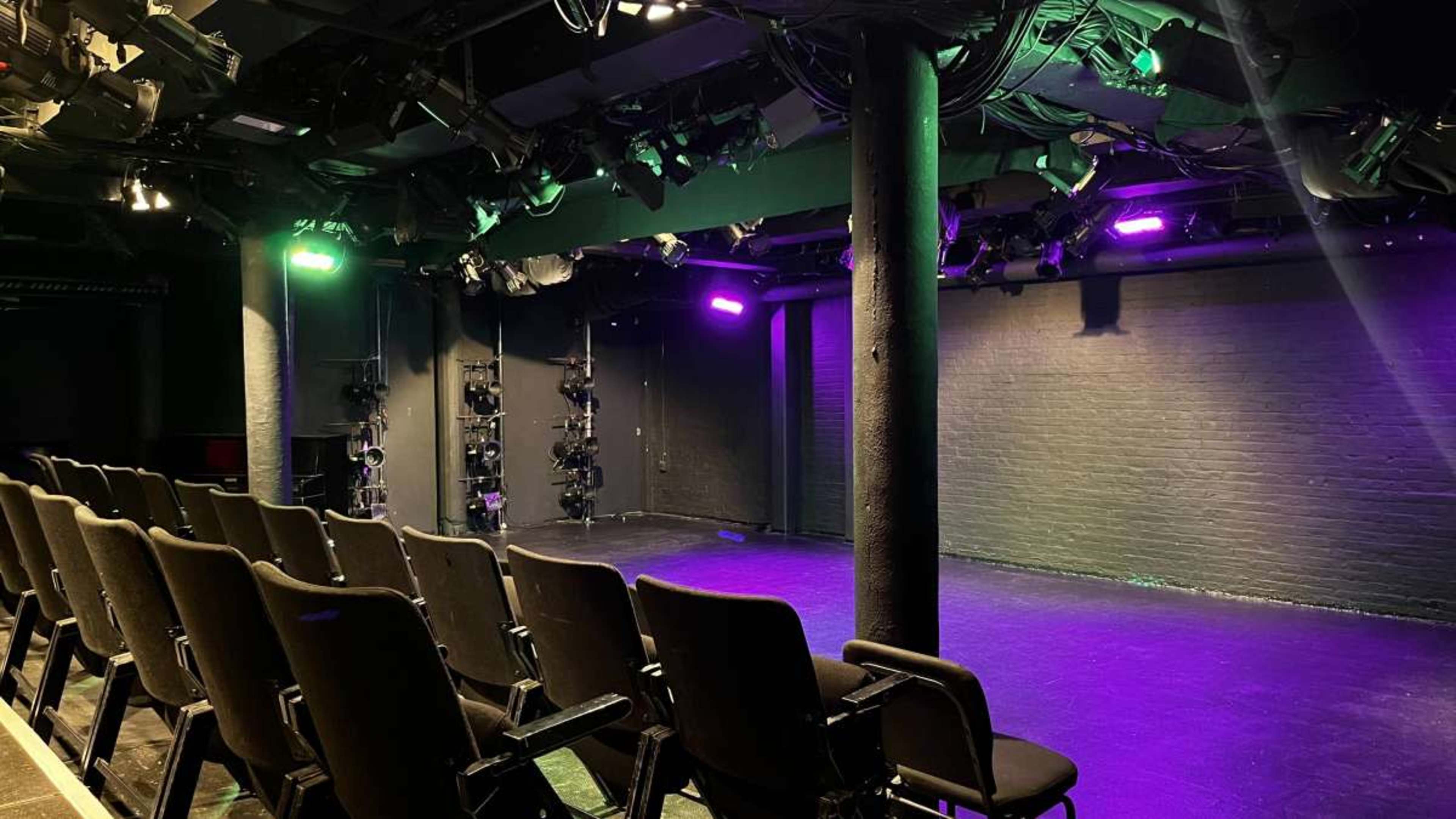 The image shows an empty theater stage with rows of black chairs facing a well-lit performance area featuring colorful stage lights.