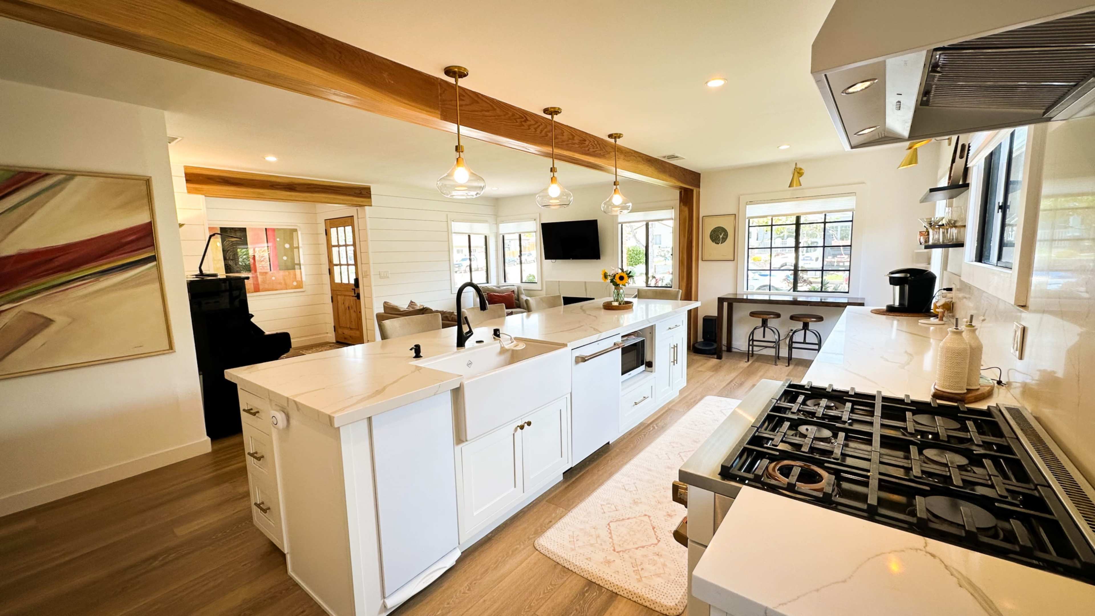 The image shows a modern kitchen with an island, a gas stove, and a dining area featuring bar stools and natural light from large windows.