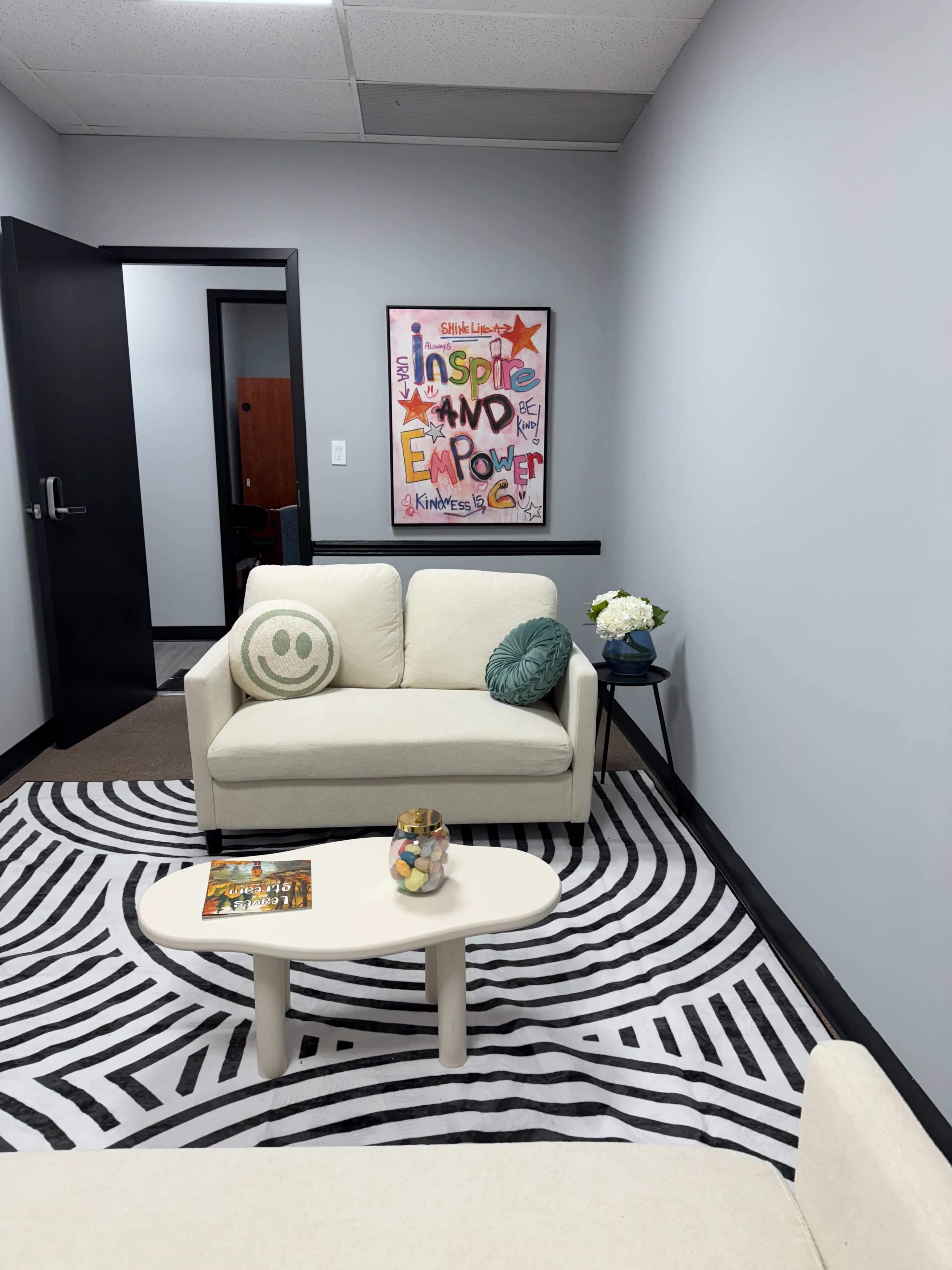 The image shows a small office space with a couch, a coffee table, a patterned rug, and a colorful wall art piece.