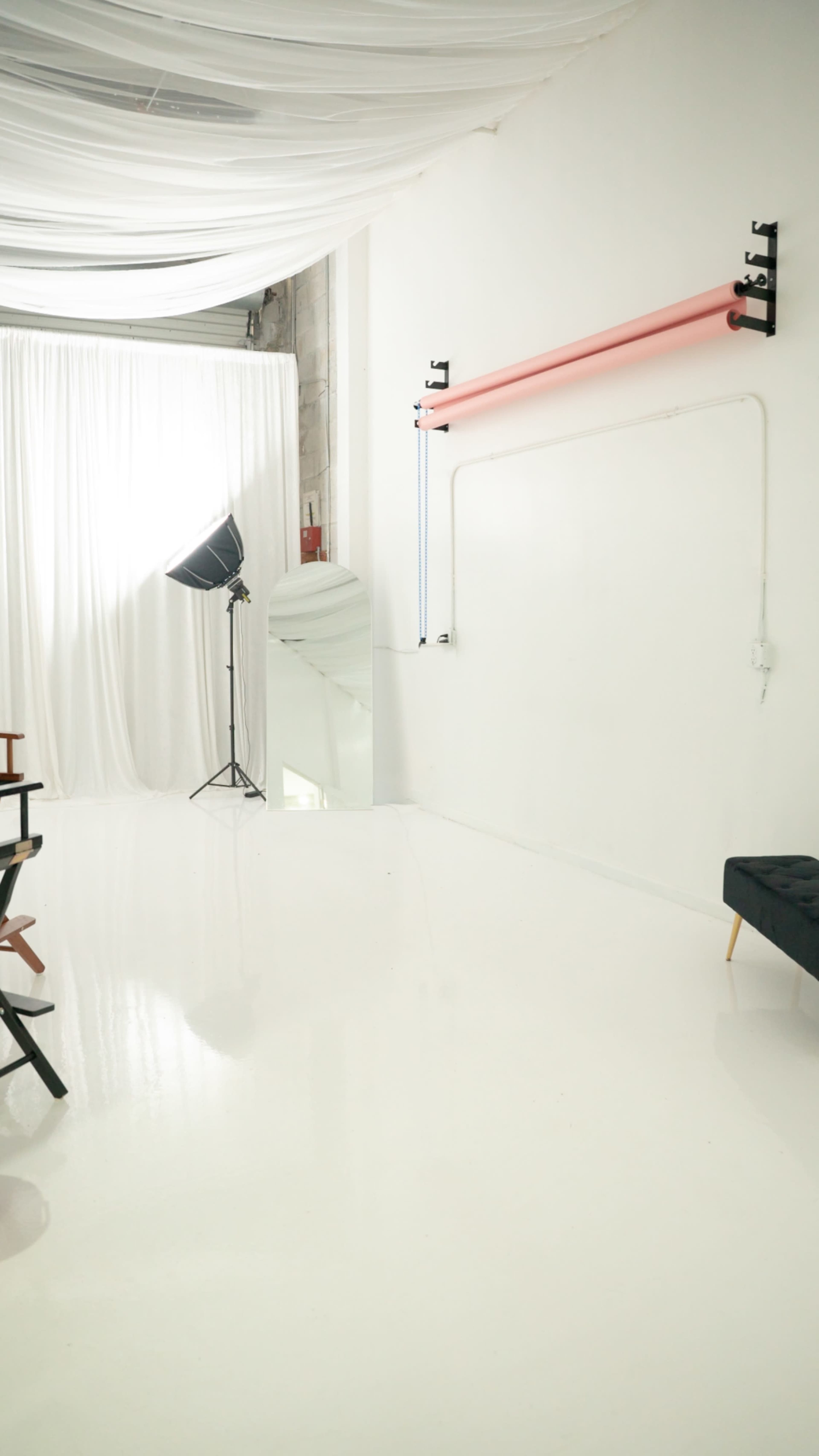 The image shows a minimalist photography studio with white walls, a mirror, a lighting setup, and a black seating area.
