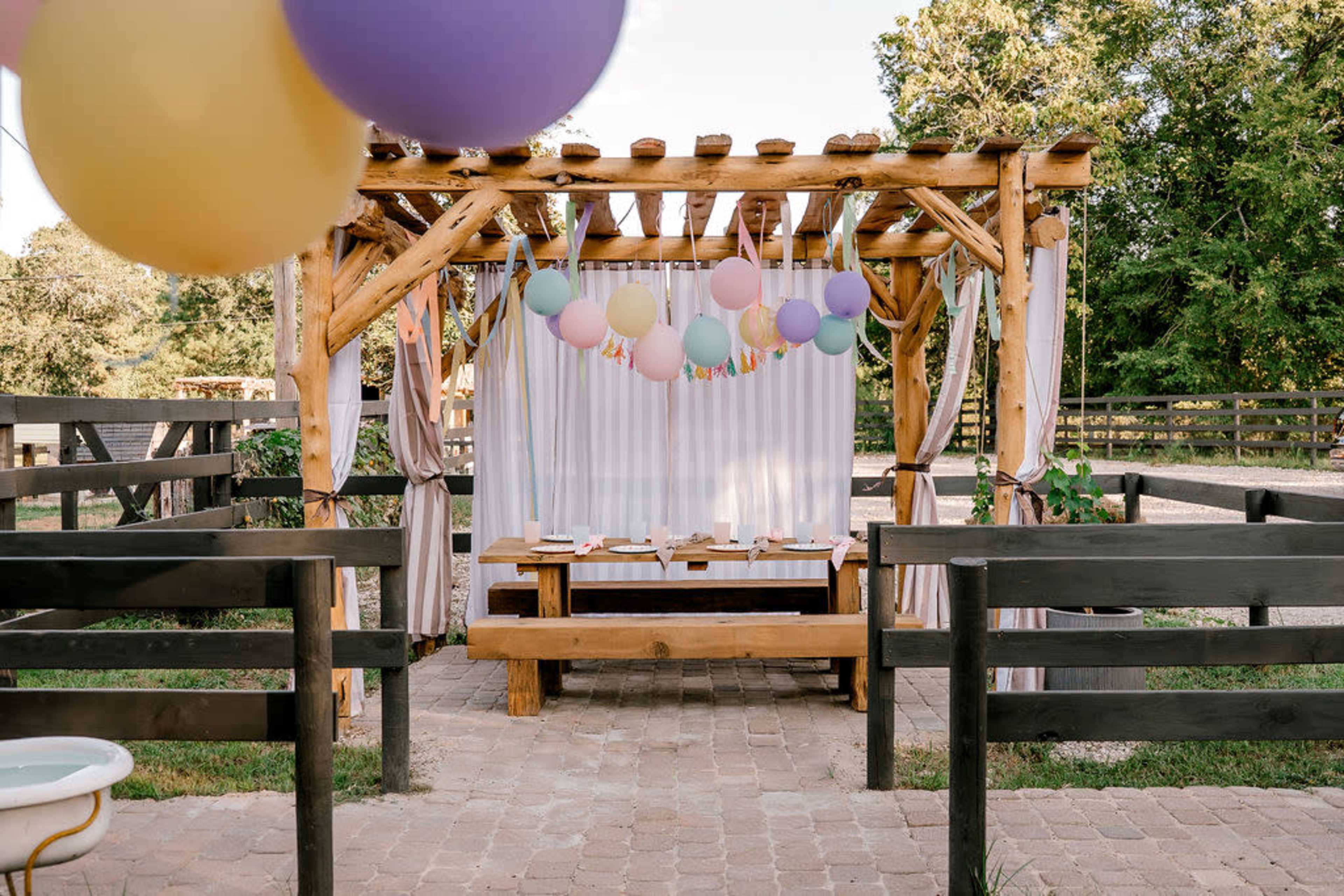 The image shows a decorated outdoor seating area with a wooden pergola, featuring balloons and a dining table set with plates and decorations.