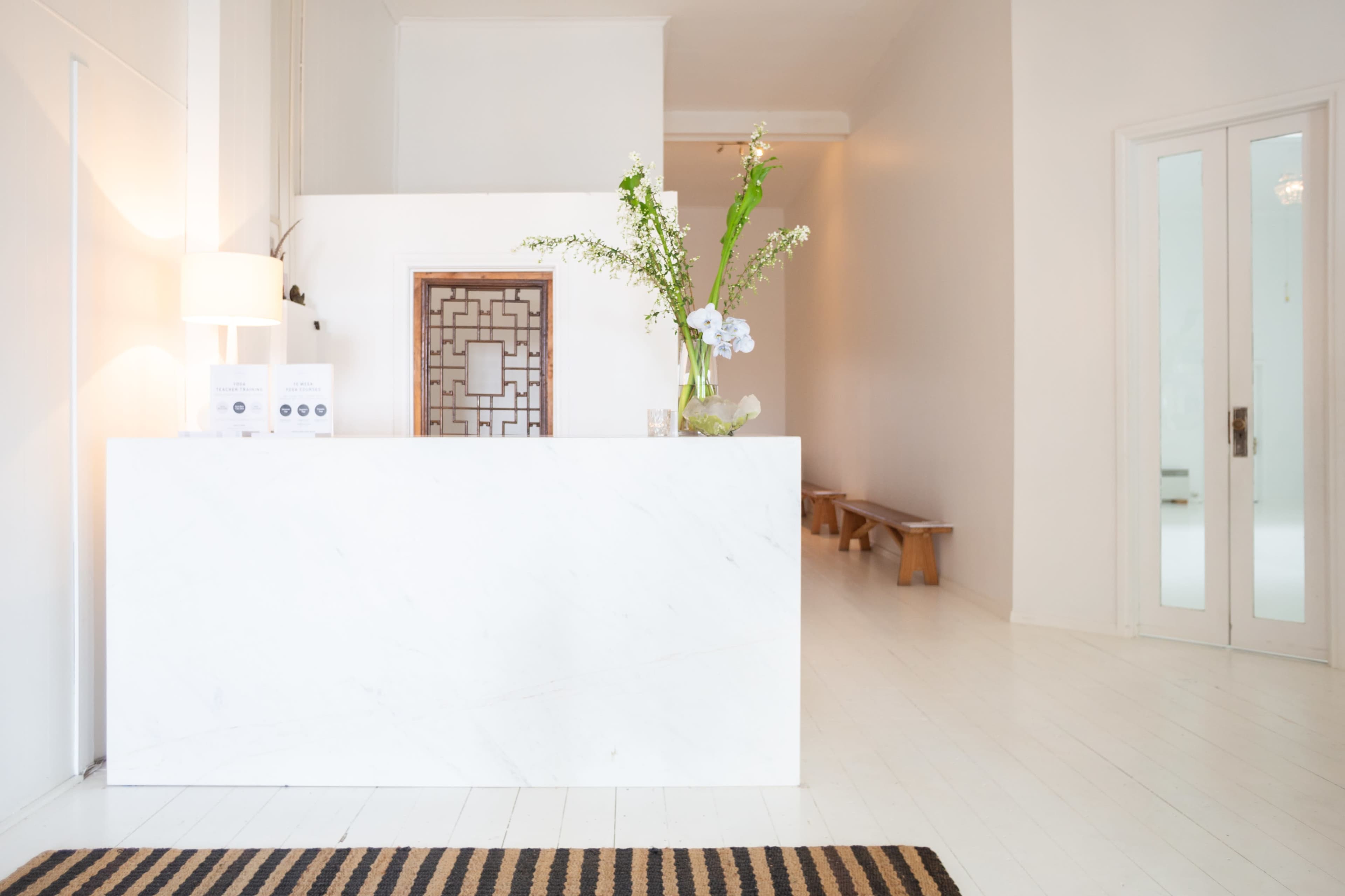 A modern reception area with a white marble front desk, a decorative mirror, and a minimalistic waiting area featuring a wooden bench.
