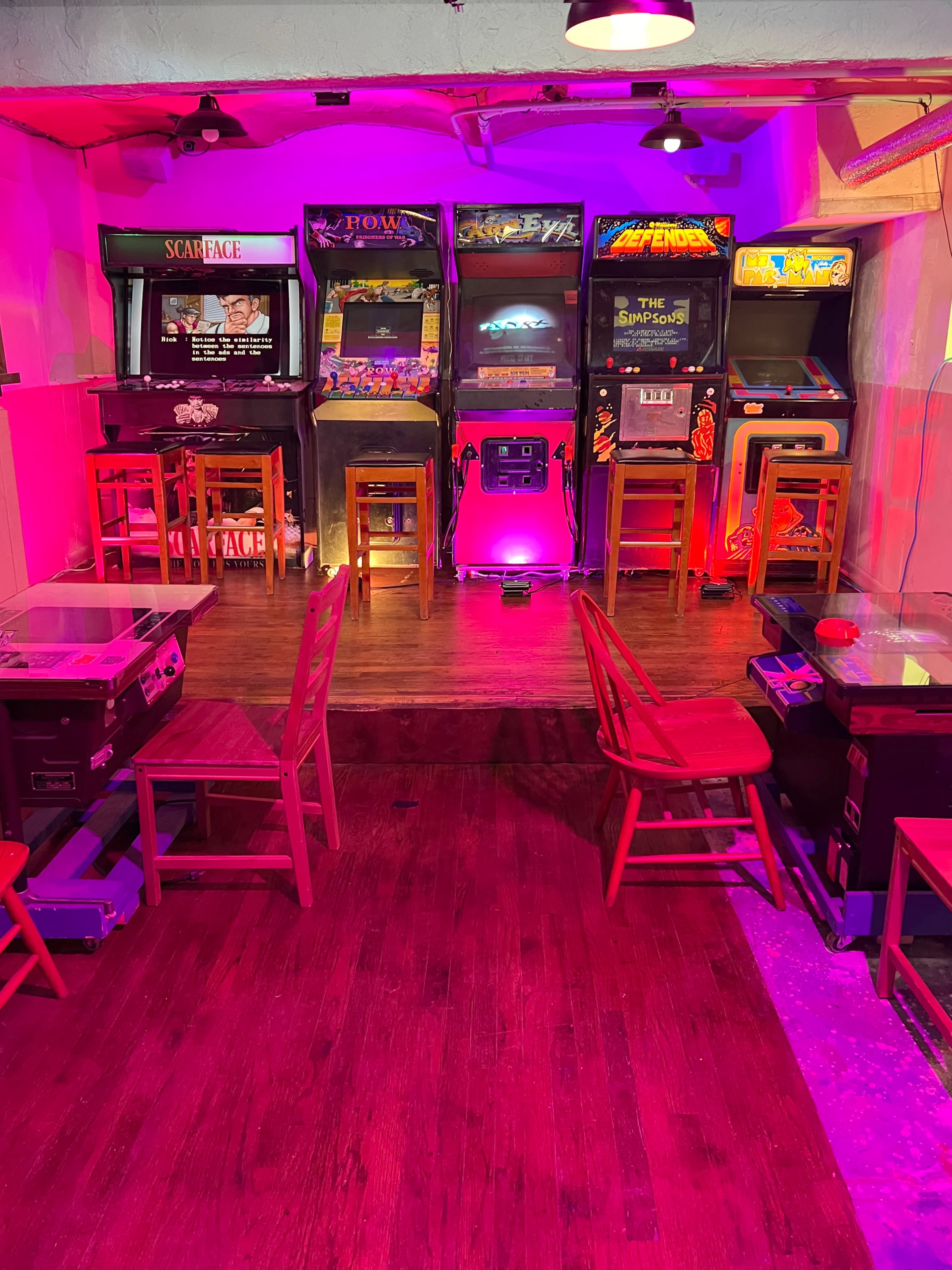 A retro arcade with several classic arcade game machines lined up against a vibrant lit wall, accompanied by wooden stools and tables.