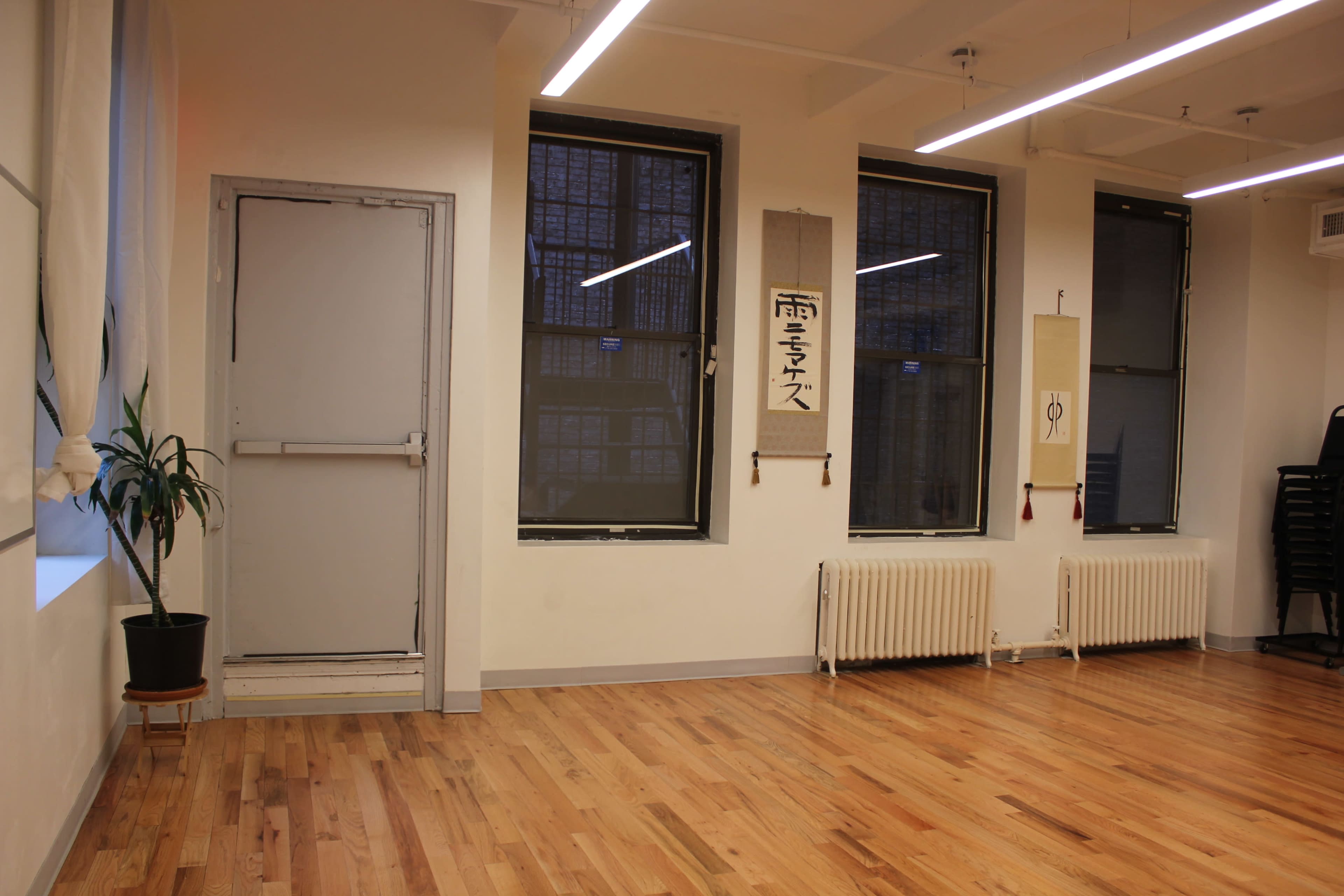 A minimalist interior of a room with wooden flooring, two windows, a door, a plant in a pot, and wall hangings.