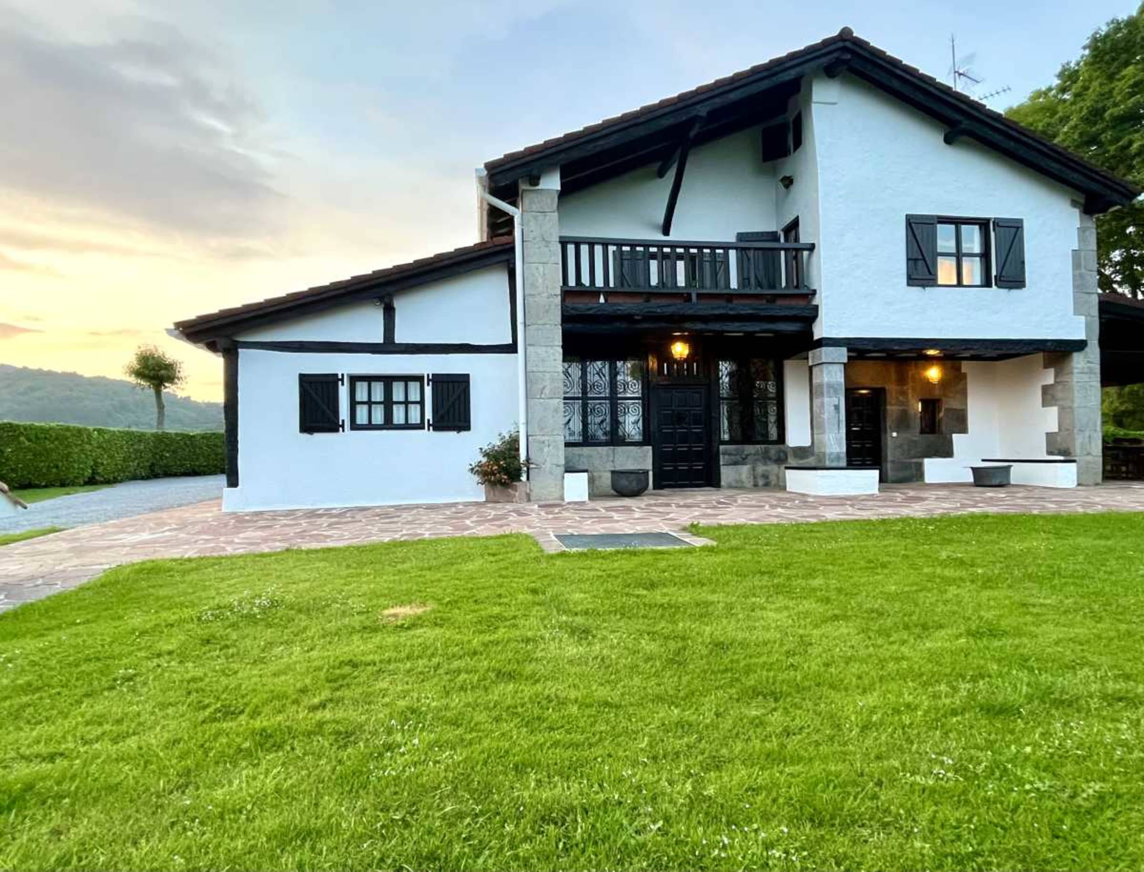 A two-story house with a white exterior and black accents is surrounded by a green lawn and features a stone entrance and balcony.