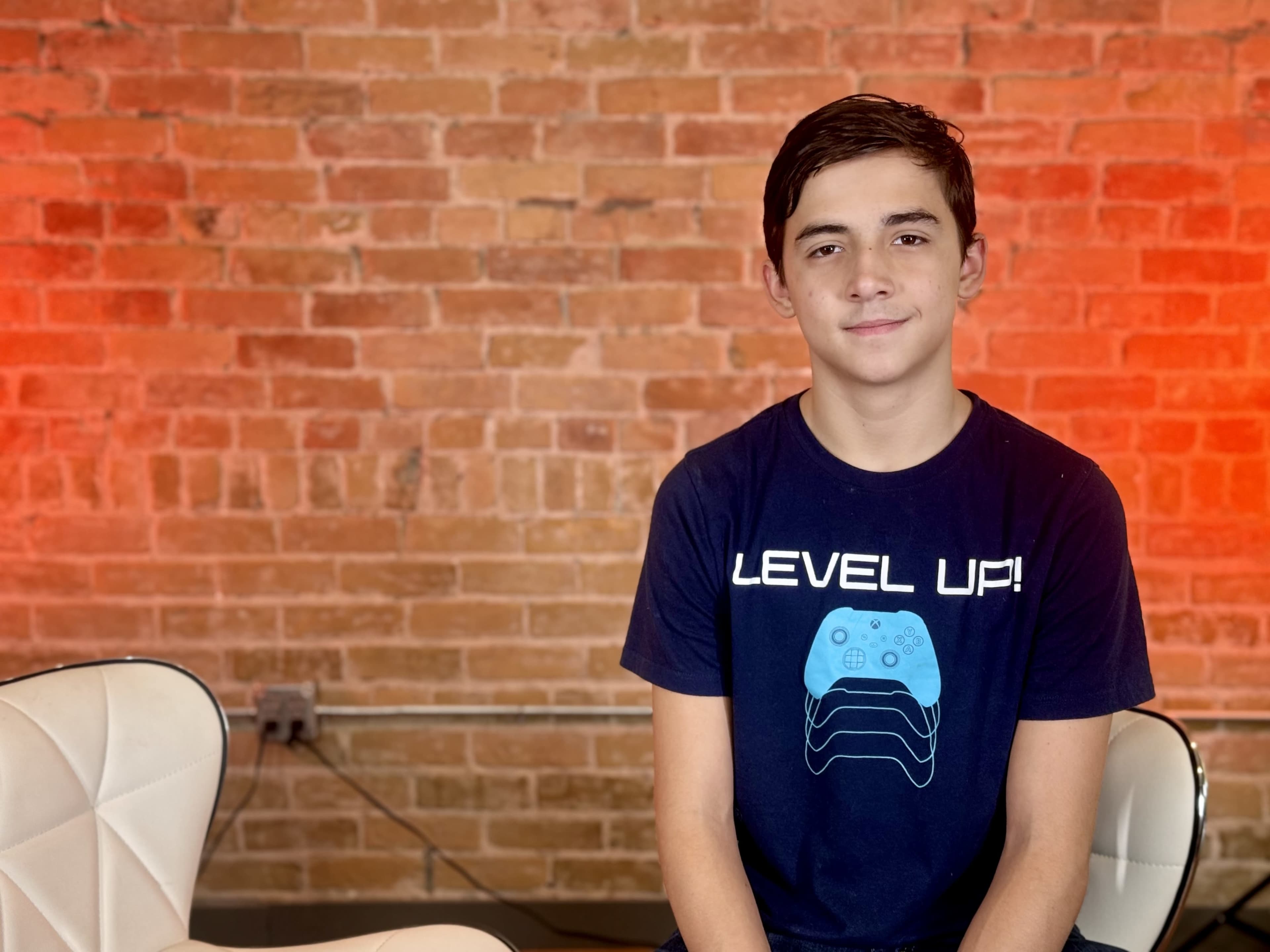 A young boy is sitting on a chair in front of a brick wall, wearing a t-shirt that says "LEVEL UP!" with a graphic of a game controller.