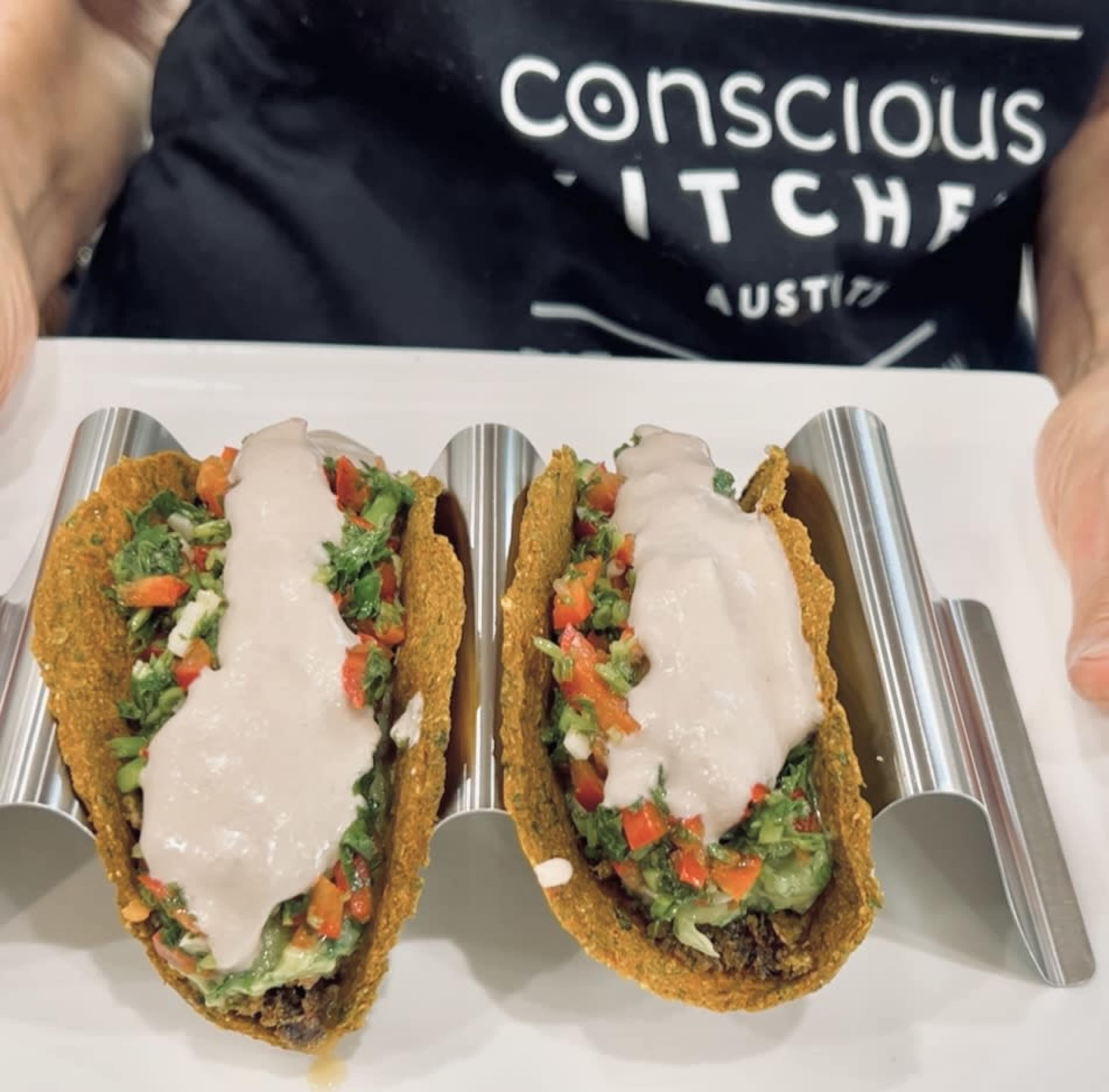 Two taco shells filled with a colorful mixture of vegetables and topped with a creamy sauce are held by a person wearing a black shirt that reads "conscious kitchen."