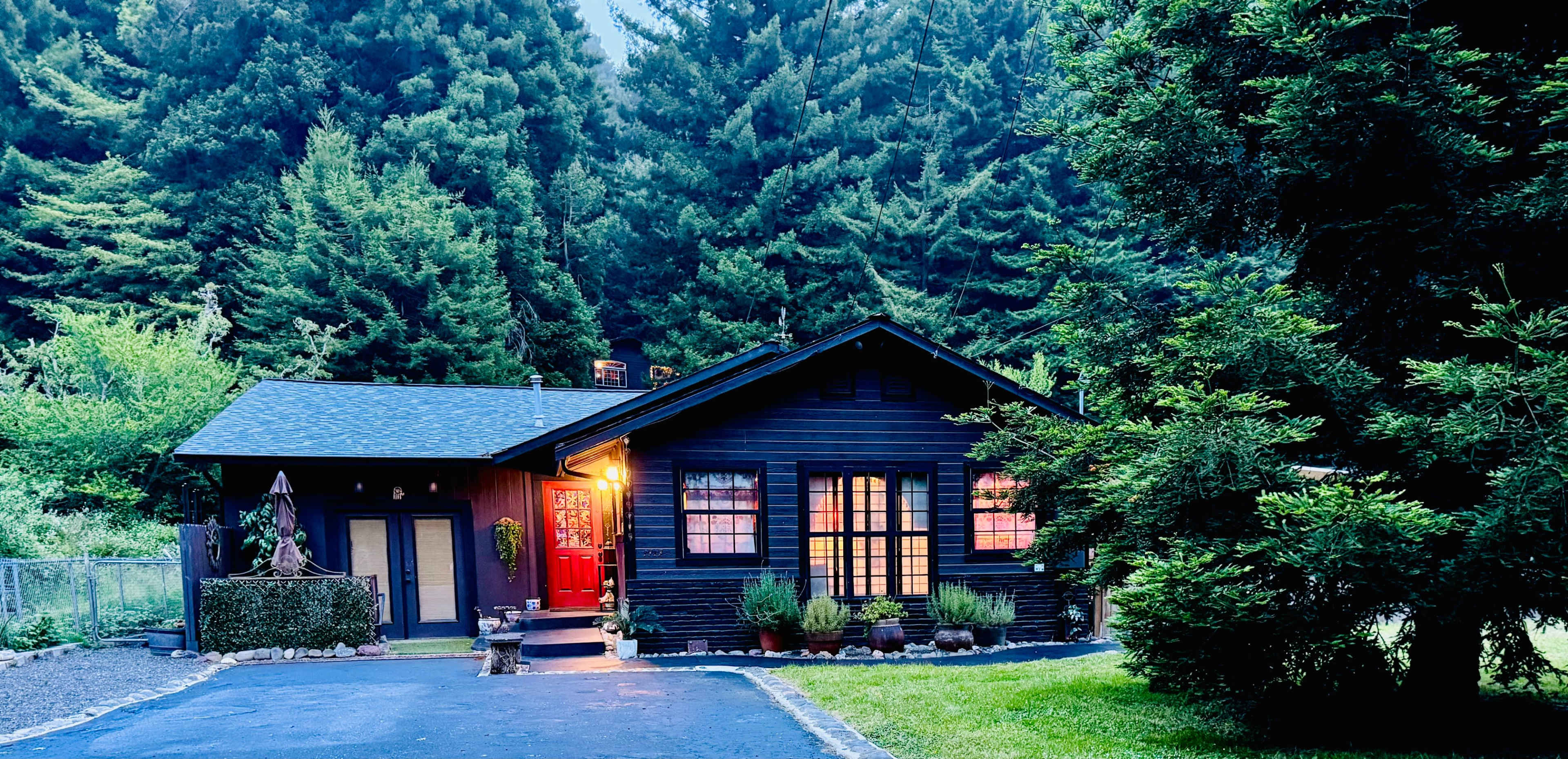 A dark-colored house with red front doors is set amidst tall green trees and a well-maintained lawn.