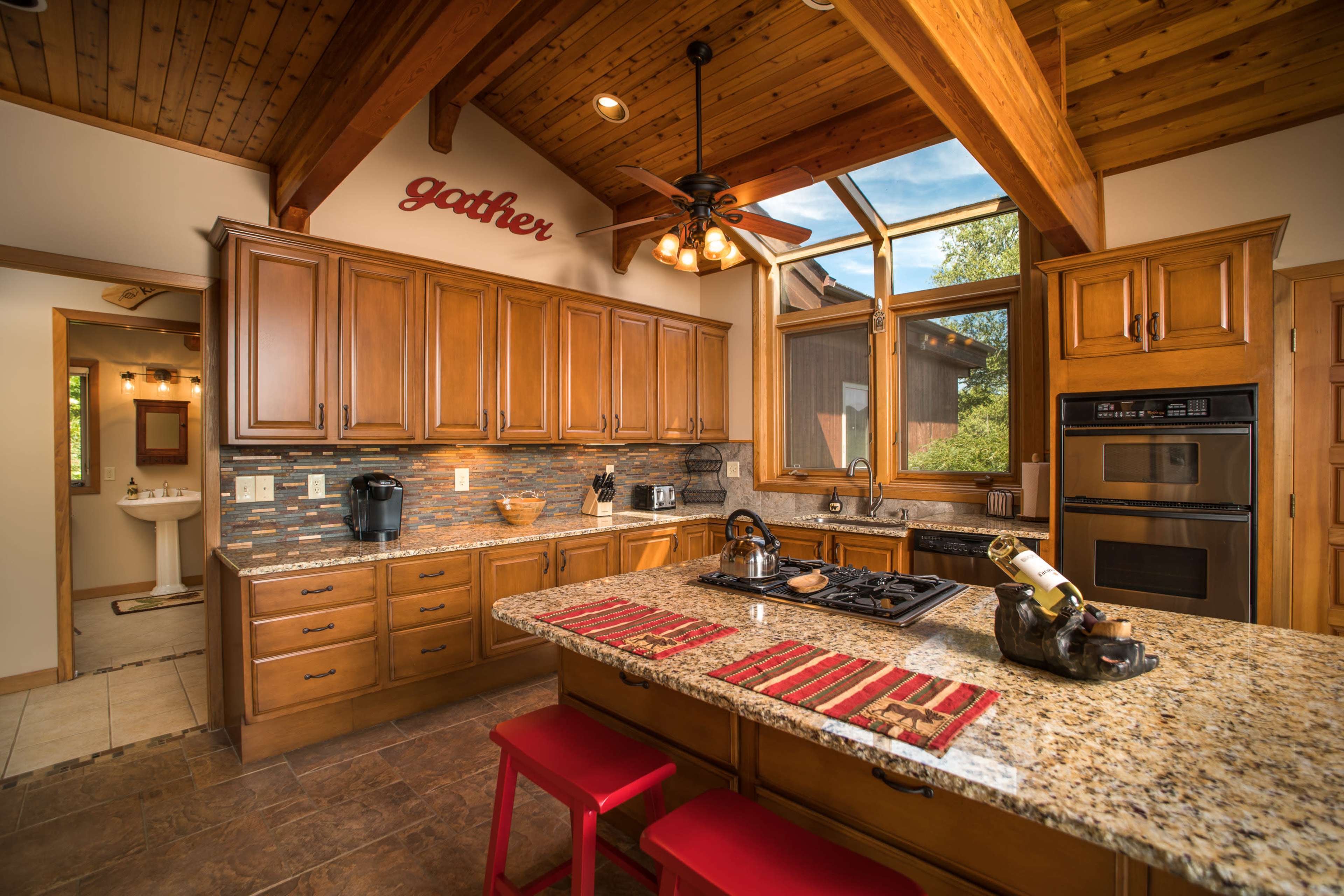 The kitchen features wooden cabinetry, a central island with red stools, and large windows that allow natural light to fill the space.