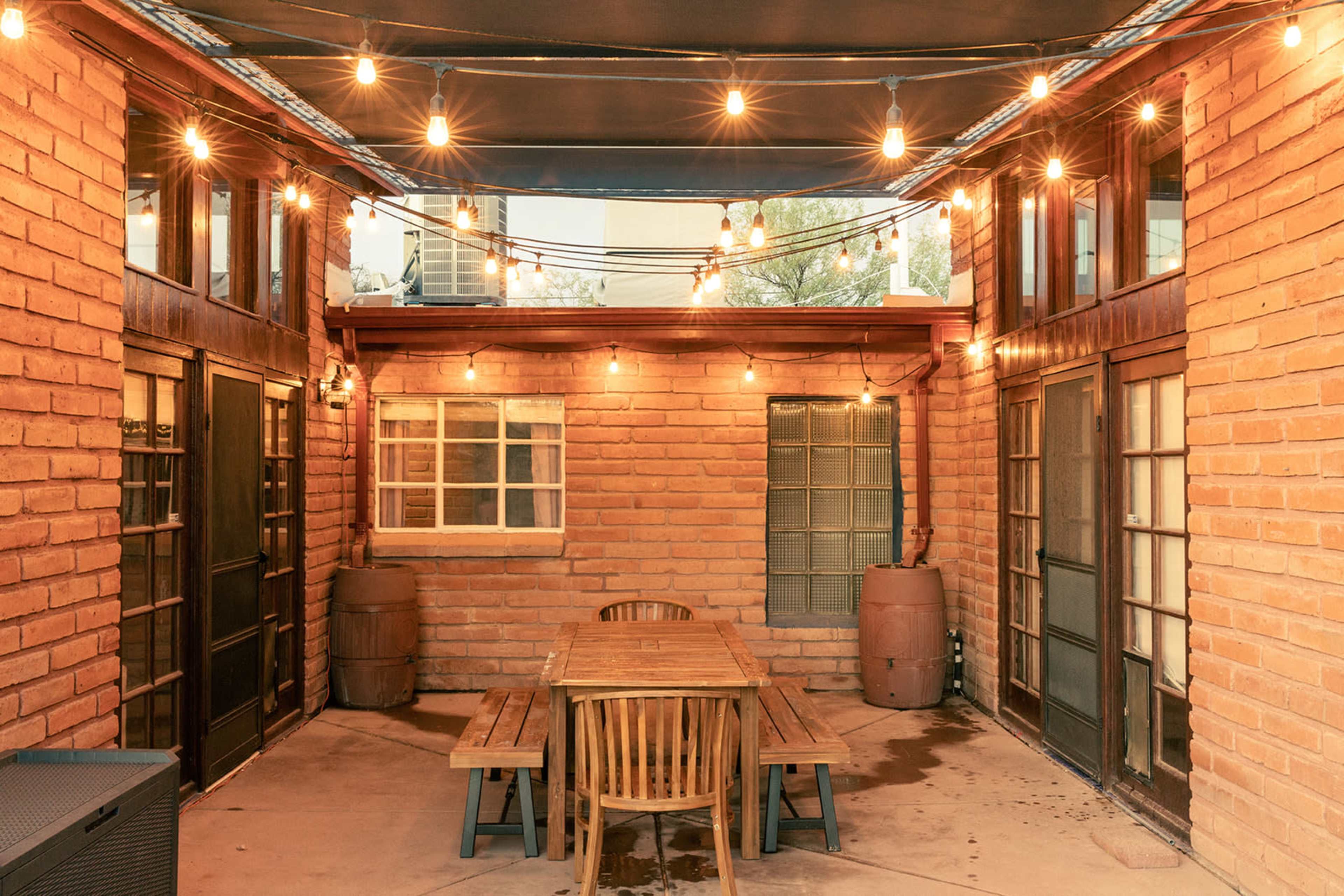 A courtyard with a wooden dining table surrounded by chairs, illuminated by string lights and brick walls.