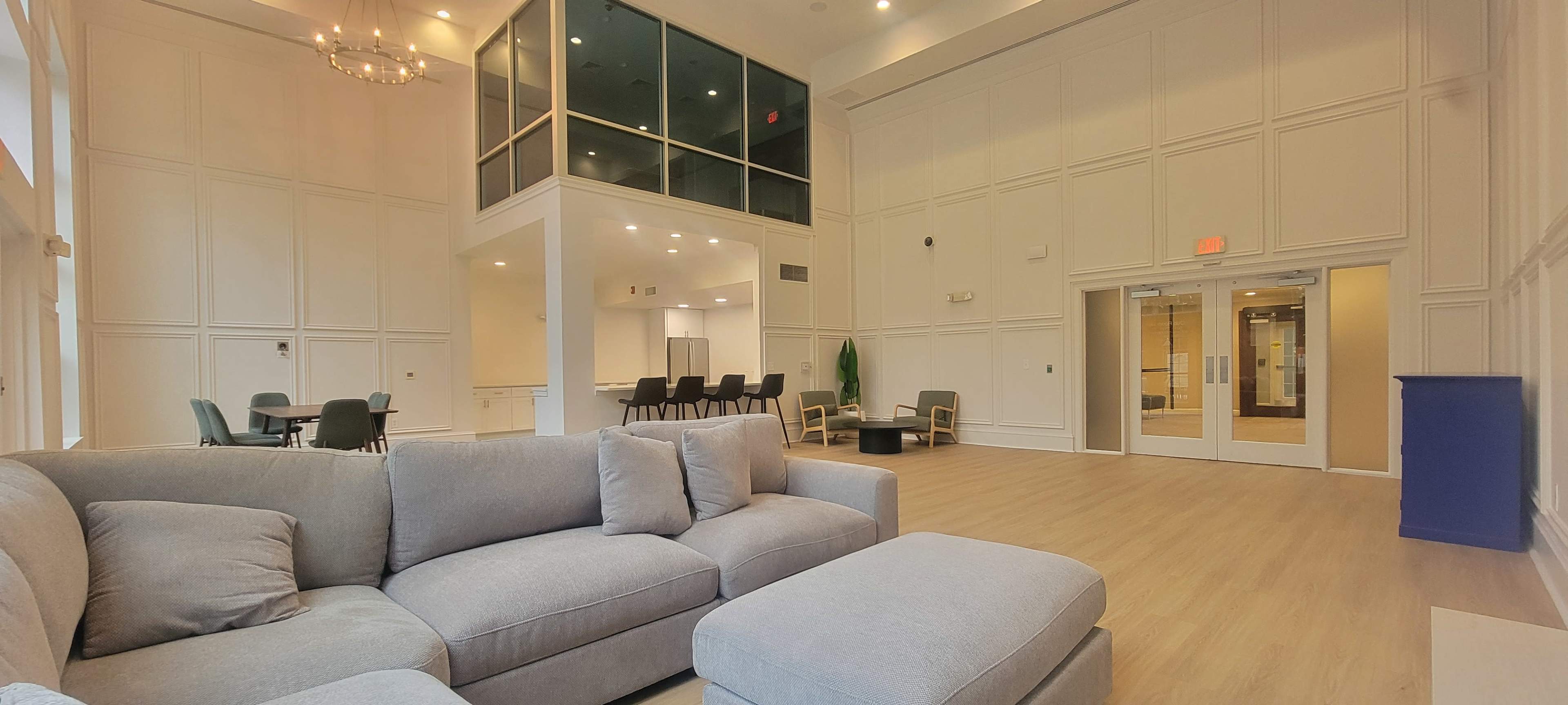 The photo shows a spacious modern lounge area with a sectional sofa, a dining table, and large windows, illuminated by natural light.