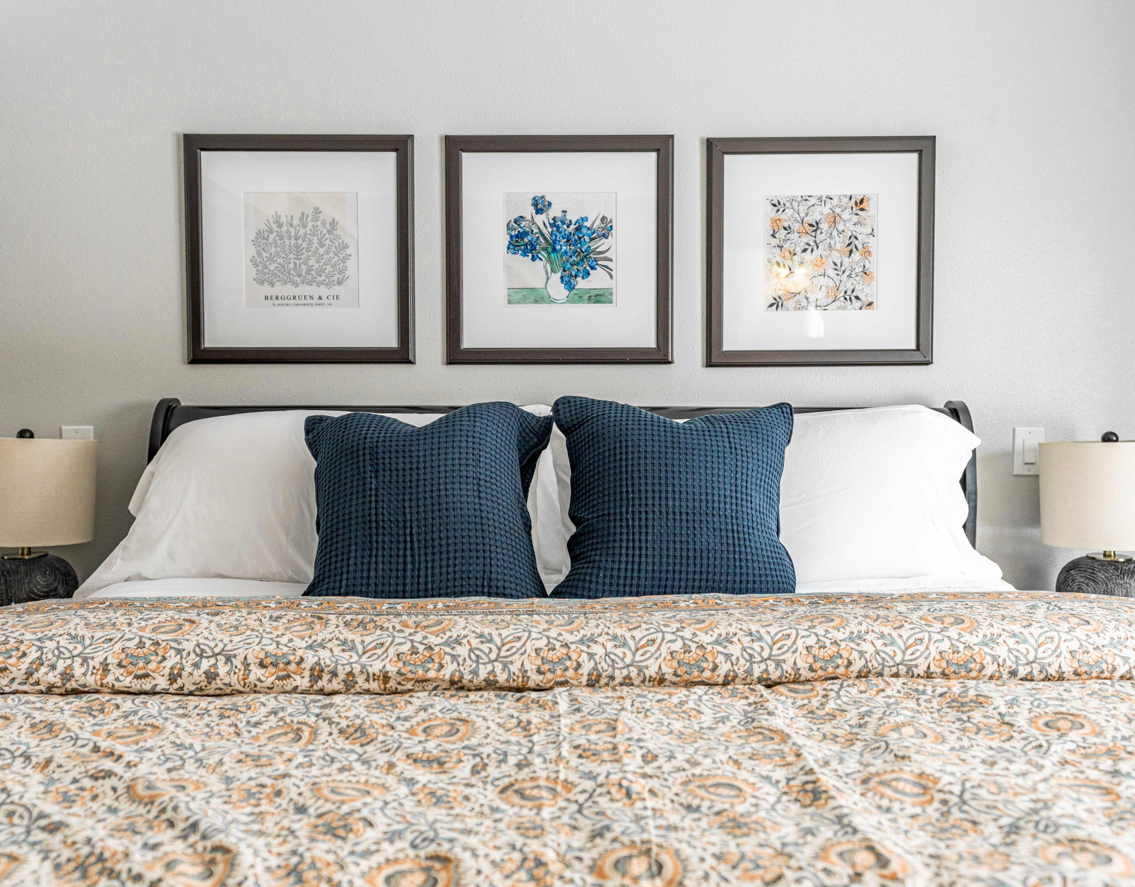 The image shows a neatly made bed with two dark blue patterned pillows, an ornate bedspread, and three framed artworks hanging on the wall above it.