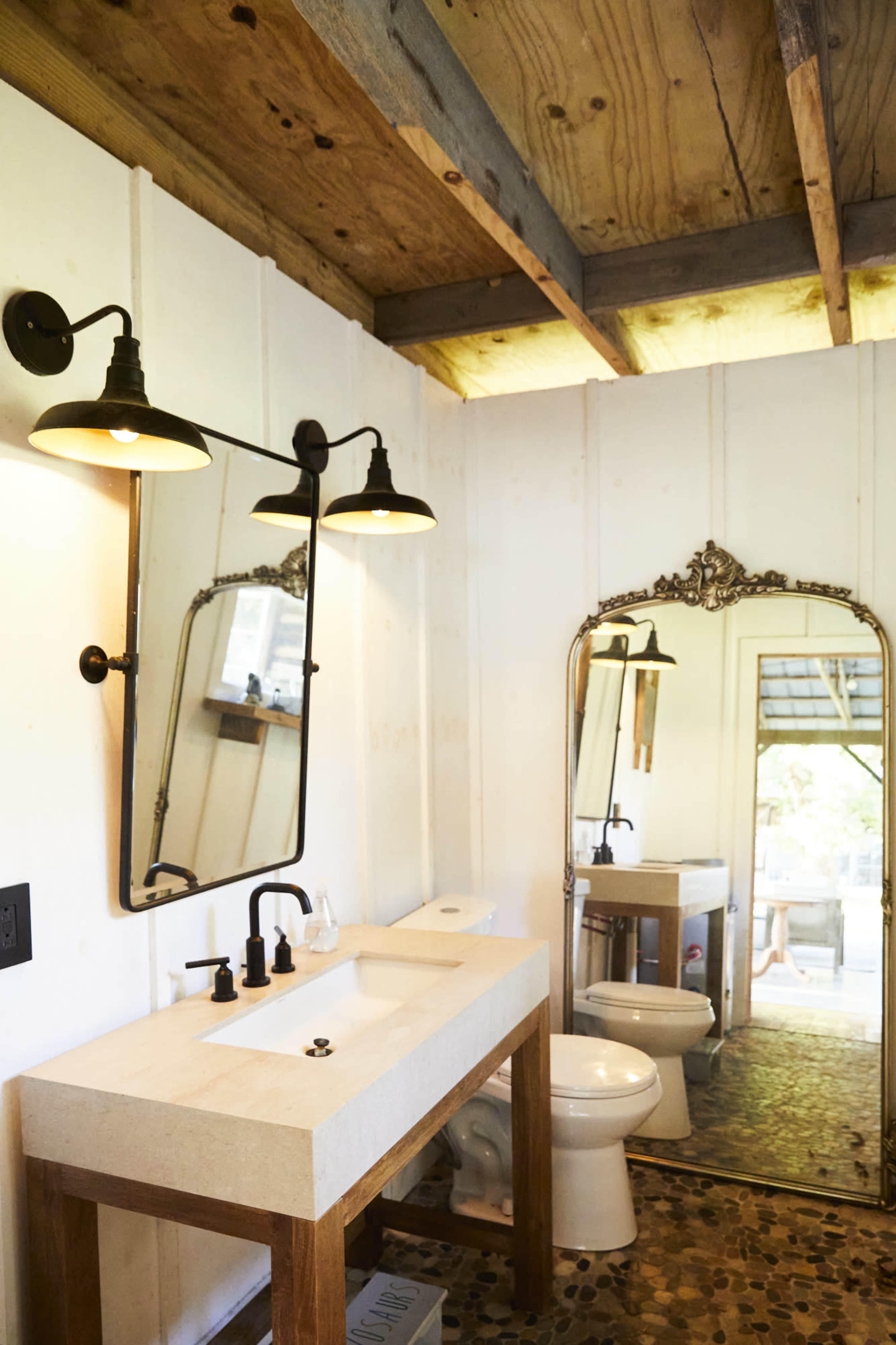 The image shows a bathroom with a wooden vanity, a rectangular sink, a wall-mounted mirror, and a vintage-style mirror, featuring a wooden ceiling and stone flooring.