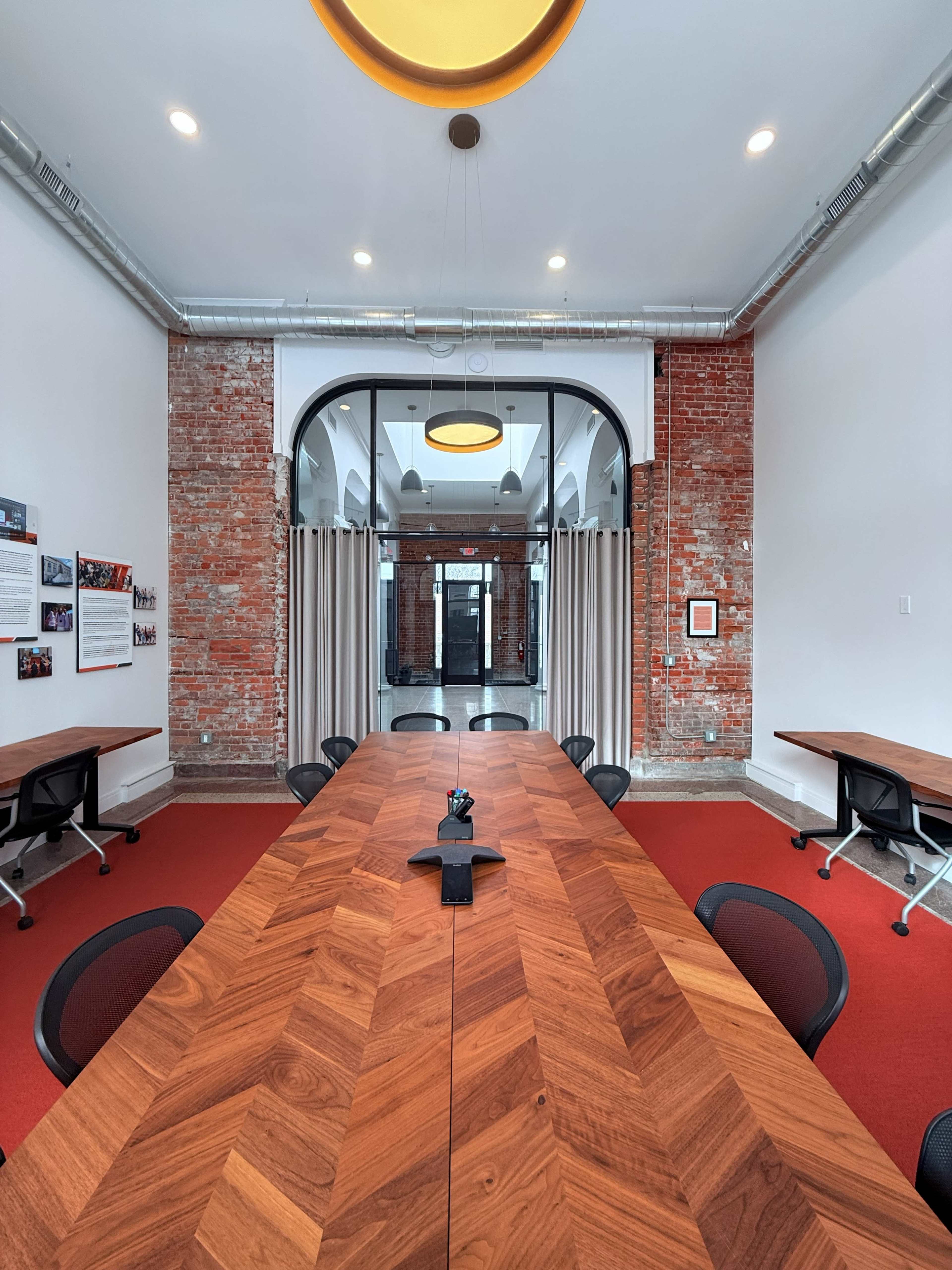 The image shows a modern conference room with a long wooden table, black chairs, exposed brick walls, and large curtains framing an entrance.