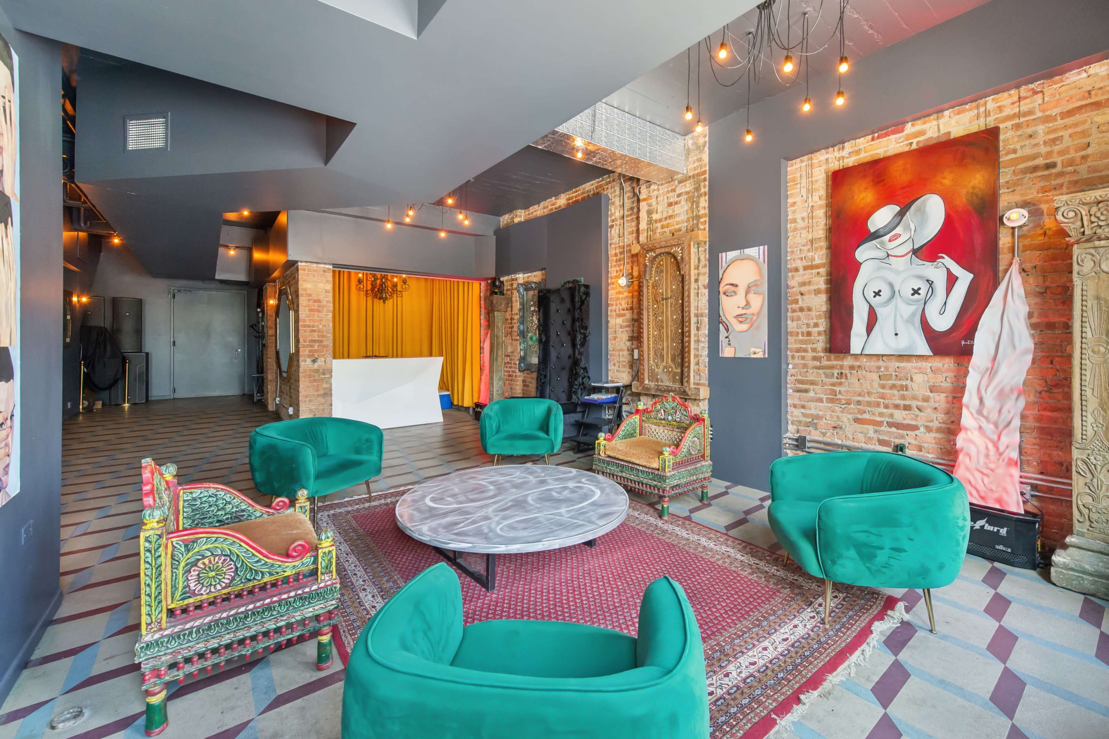 The image shows a stylish interior of a trendy venue featuring green velvet chairs arranged around a circular table, with brick walls and artistic decorations.