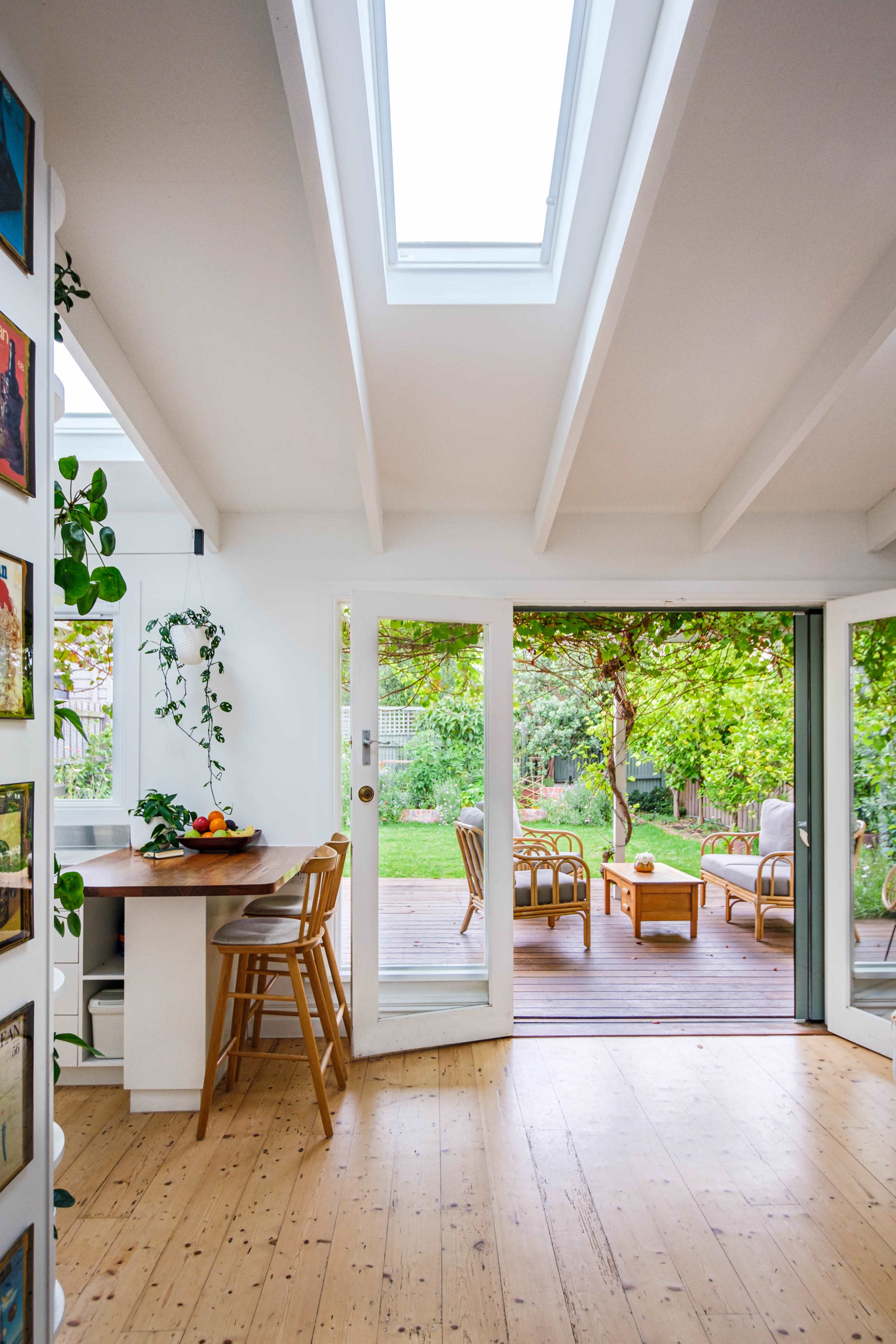 A bright, open living space features a dining area with a wooden table, surrounded by plants, leading to a deck with seating and a view of a garden.