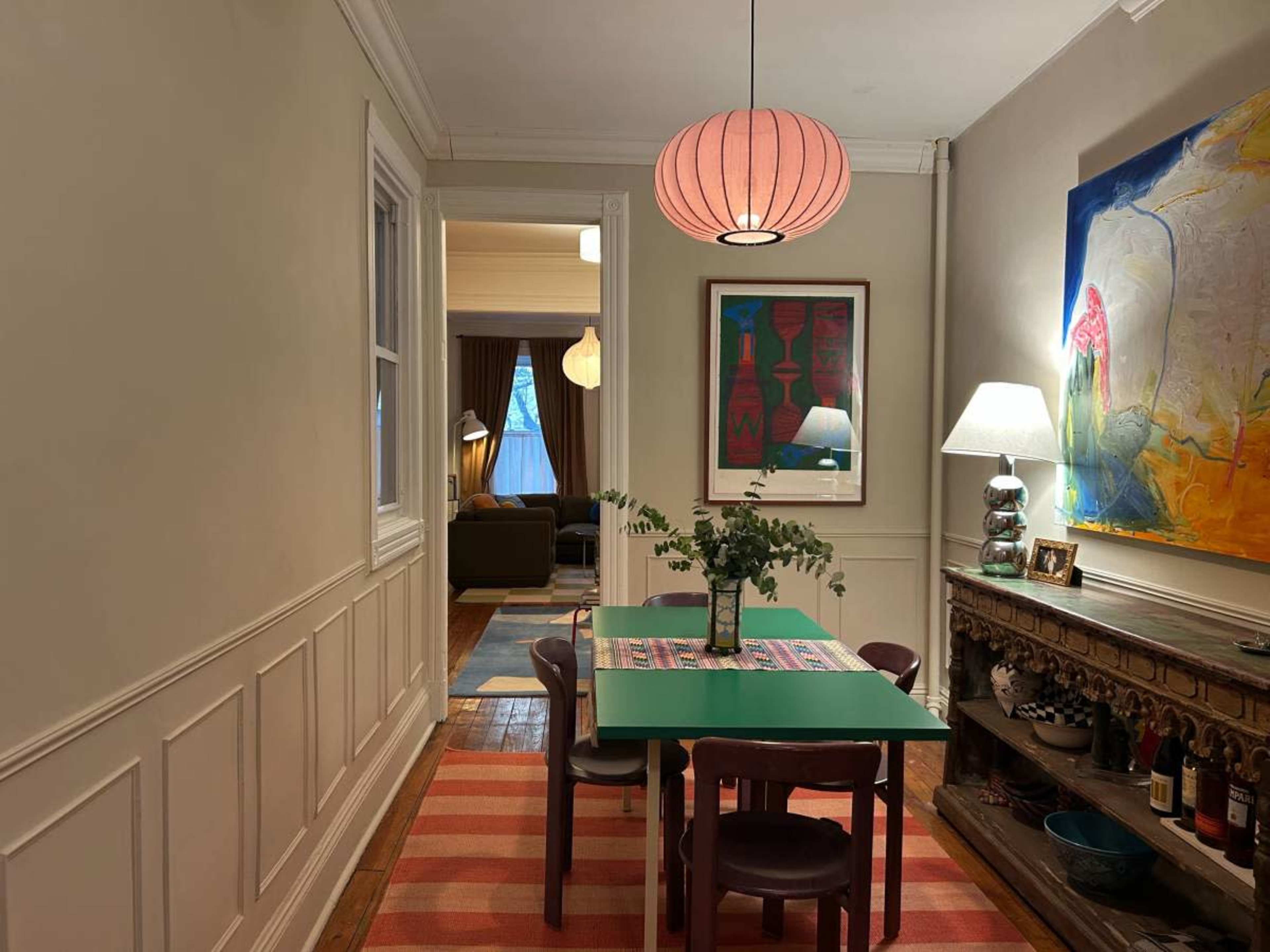The image shows a hallway with a green dining table surrounded by chairs, a decorative lamp, and an art piece on the wall, leading to a living room area.
