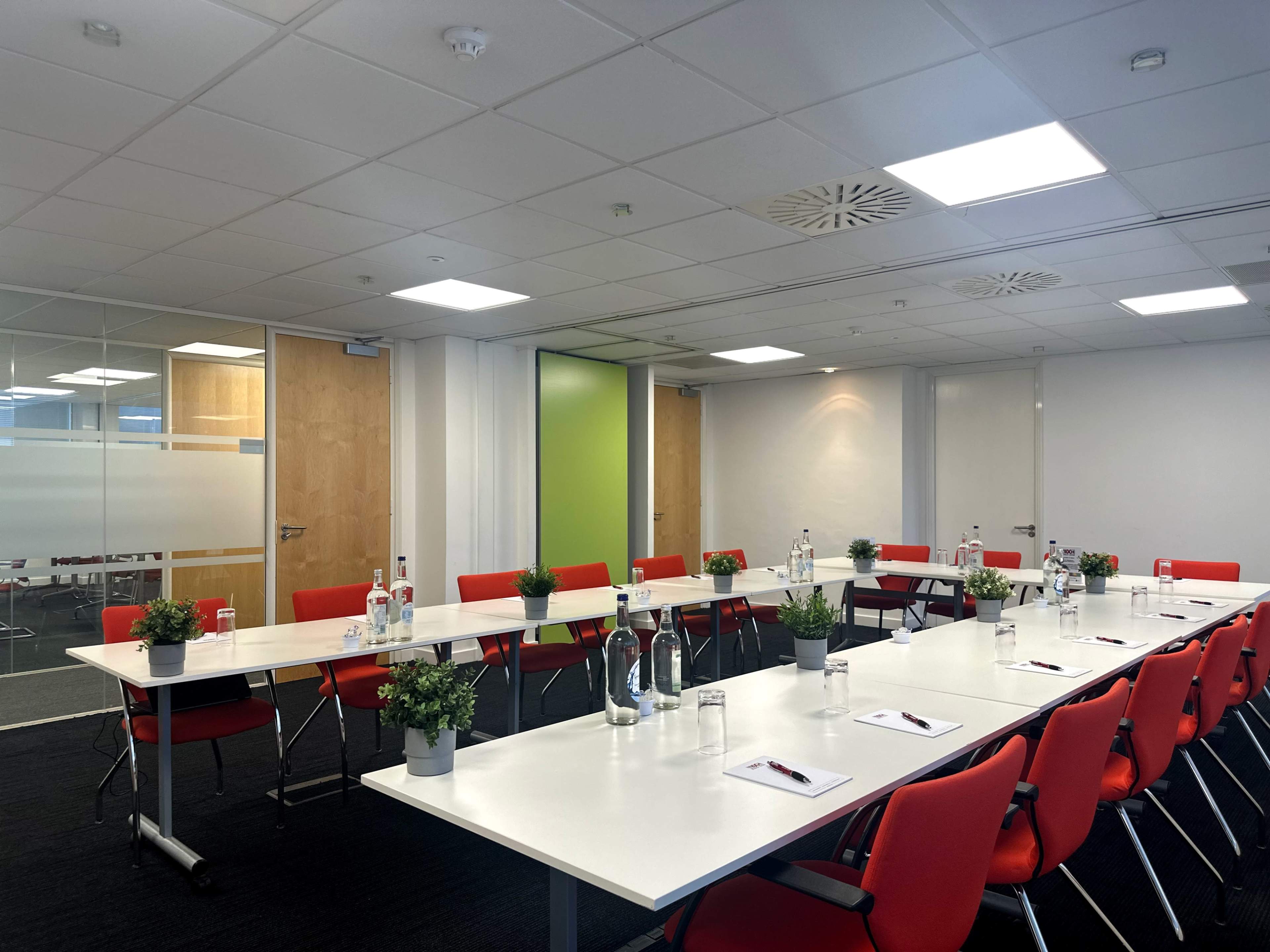 A modern conference room features a long white table with red chairs, plants, and water bottles, set up for a meeting.