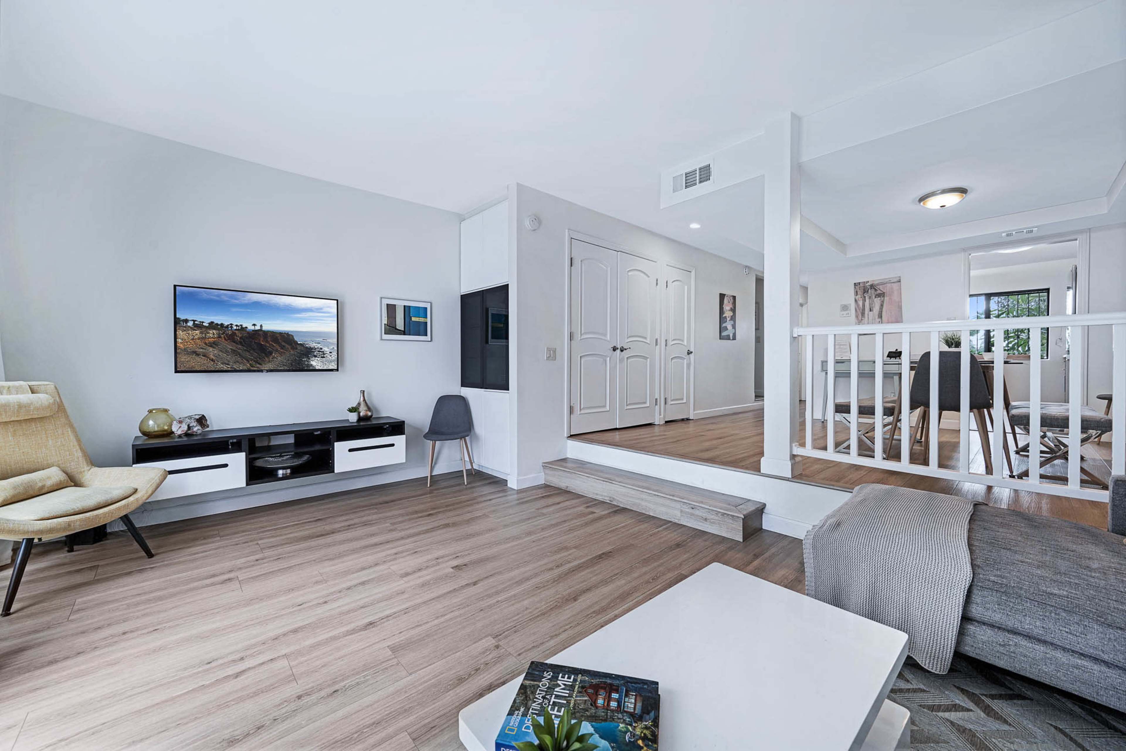 The image shows a modern living room with light wood flooring, a flat-screen TV mounted on the wall, and a small coffee table, leading to an adjacent dining area.
