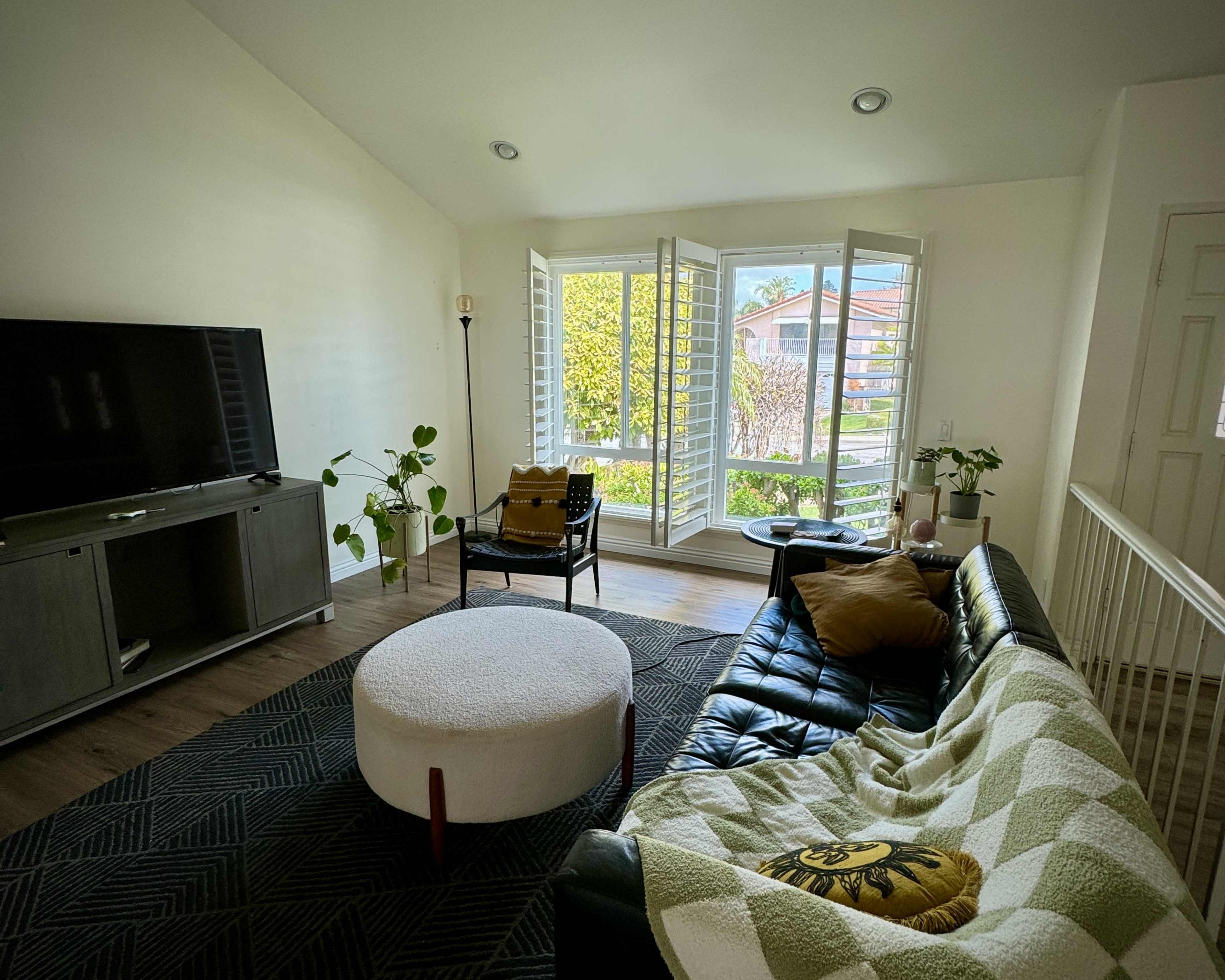 A bright living room with a large window, a black leather sofa, a round white coffee table, and various plants.
