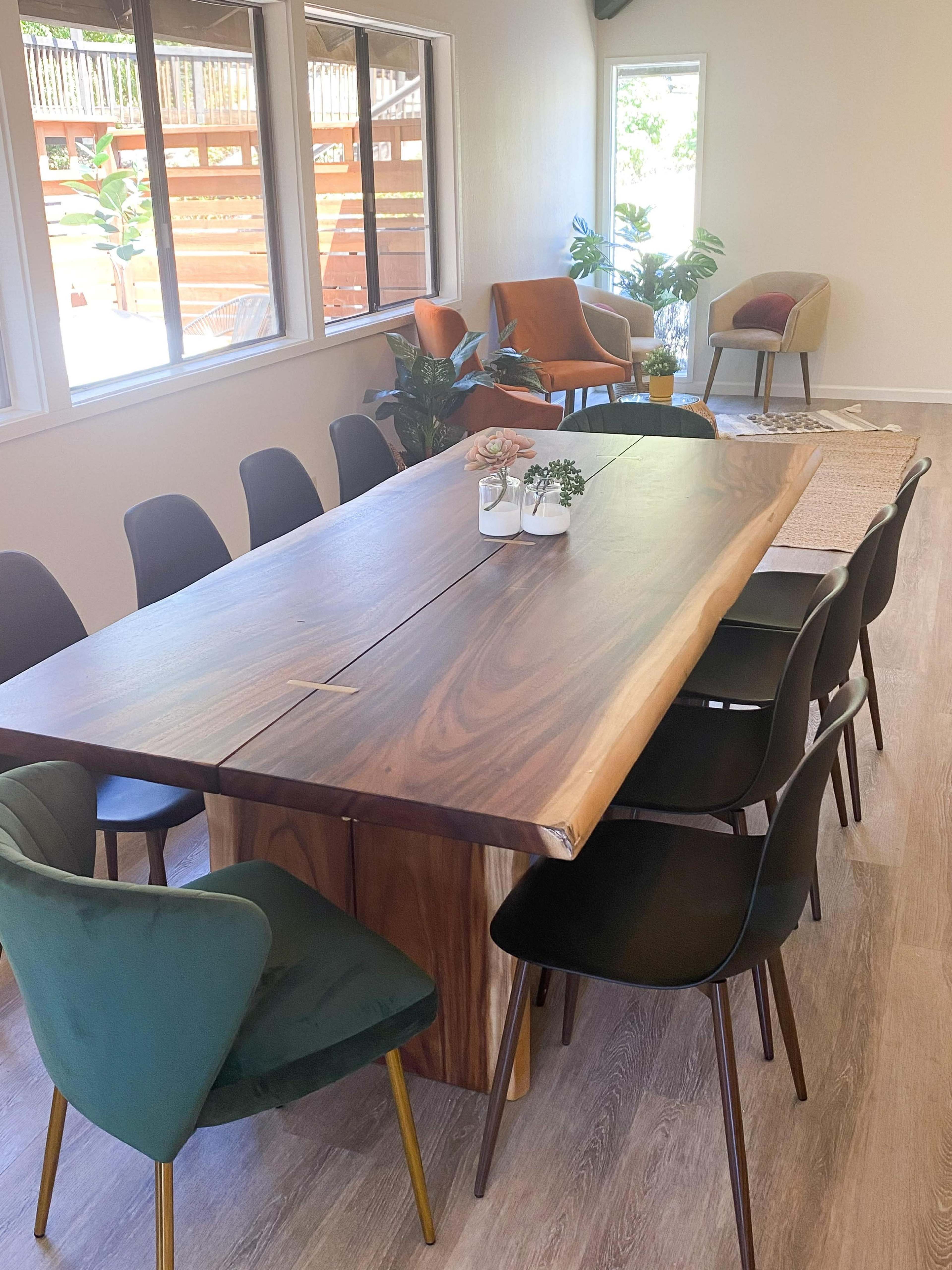 A wooden dining table surrounded by black and green chairs is set in a bright room with large windows and various indoor plants.