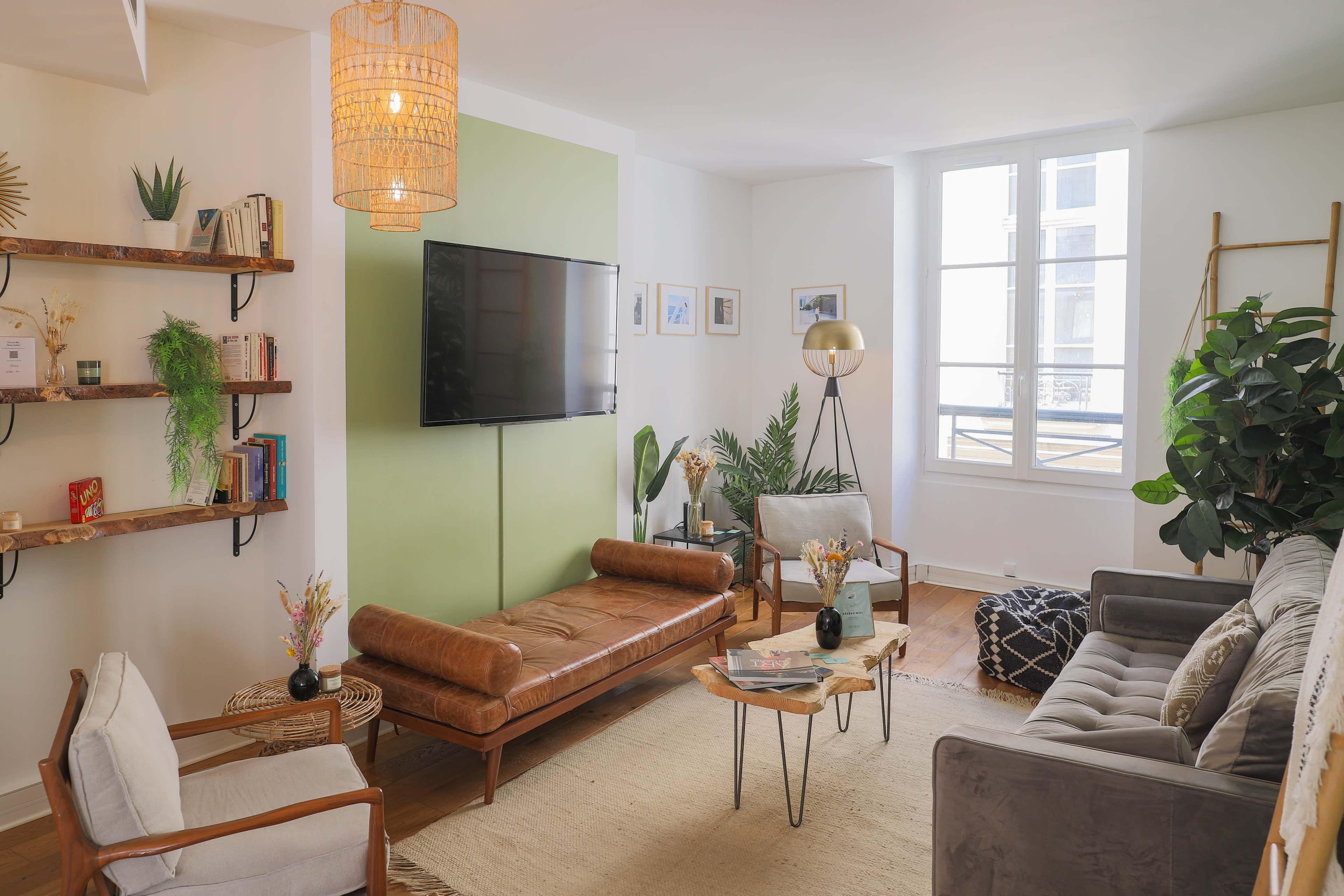 The image shows a modern living room featuring a brown leather sofa, a gray couch, a television mounted on a green accent wall, and various indoor plants arranged around a coffee table.