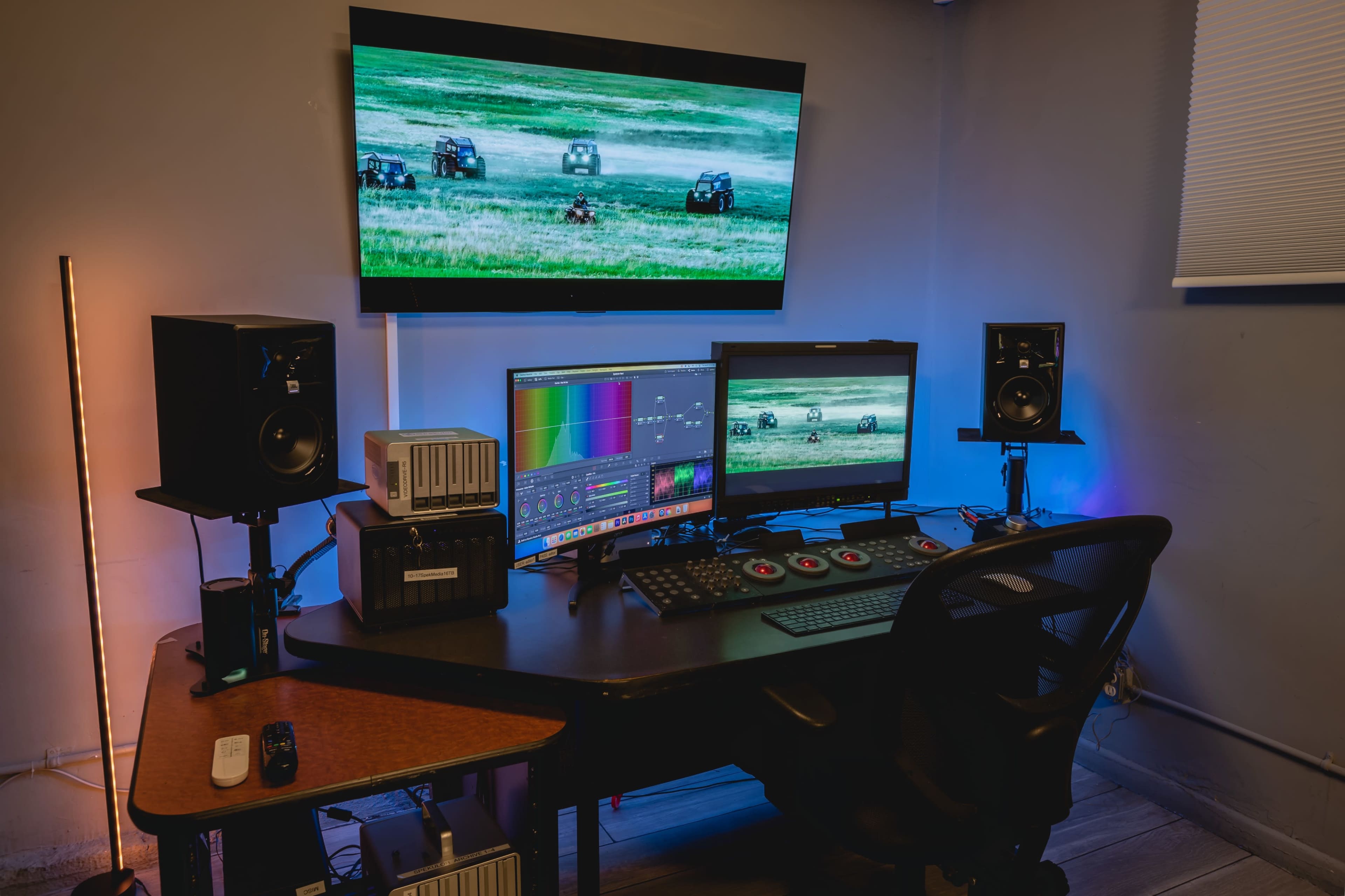 A video editing workstation with two monitors, speakers, and a control panel, set in a room with a large display showing a landscape scene.