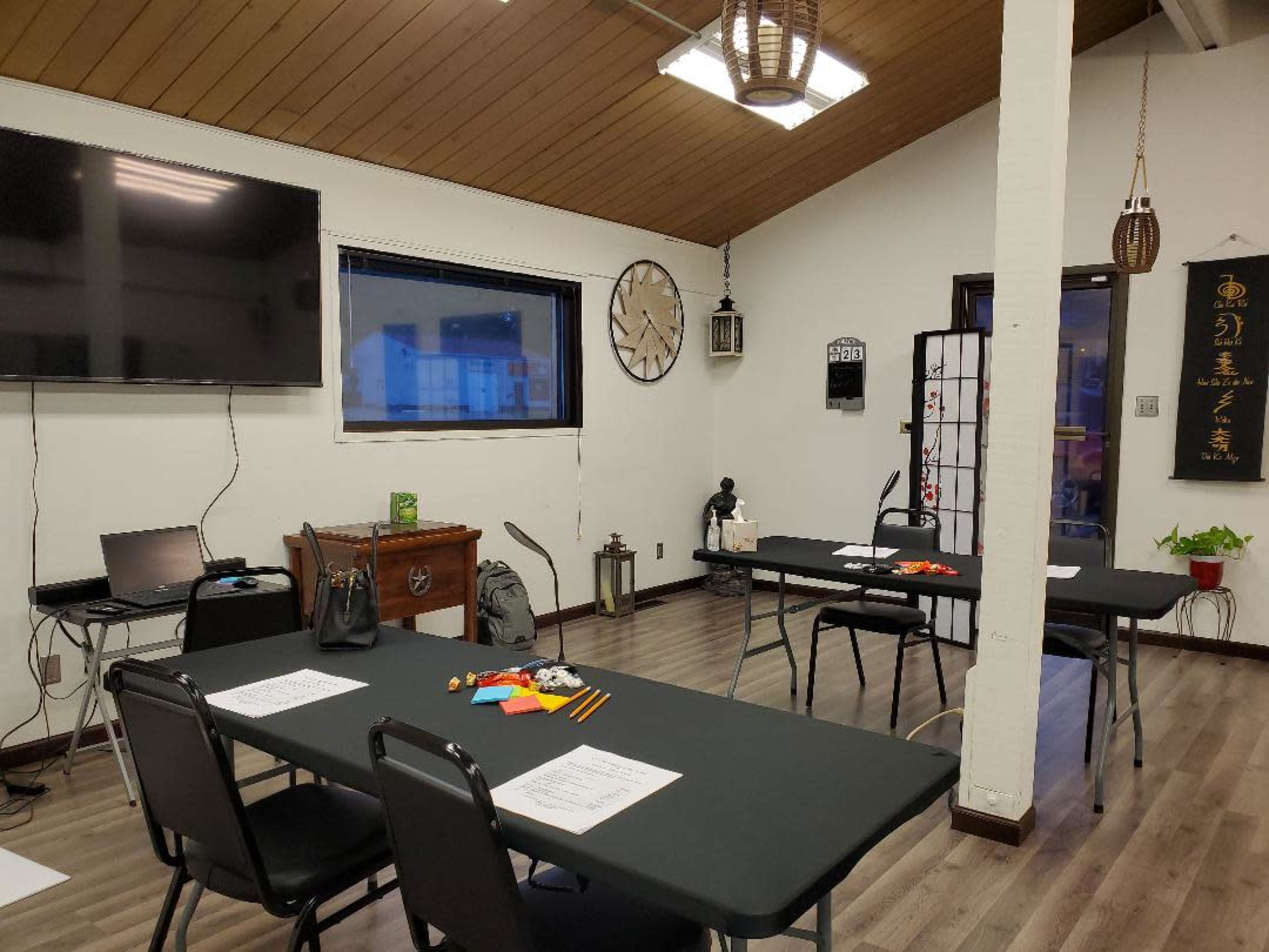 A room with several tables and chairs arranged for a meeting, featuring a television mounted on the wall and a clock above a window.