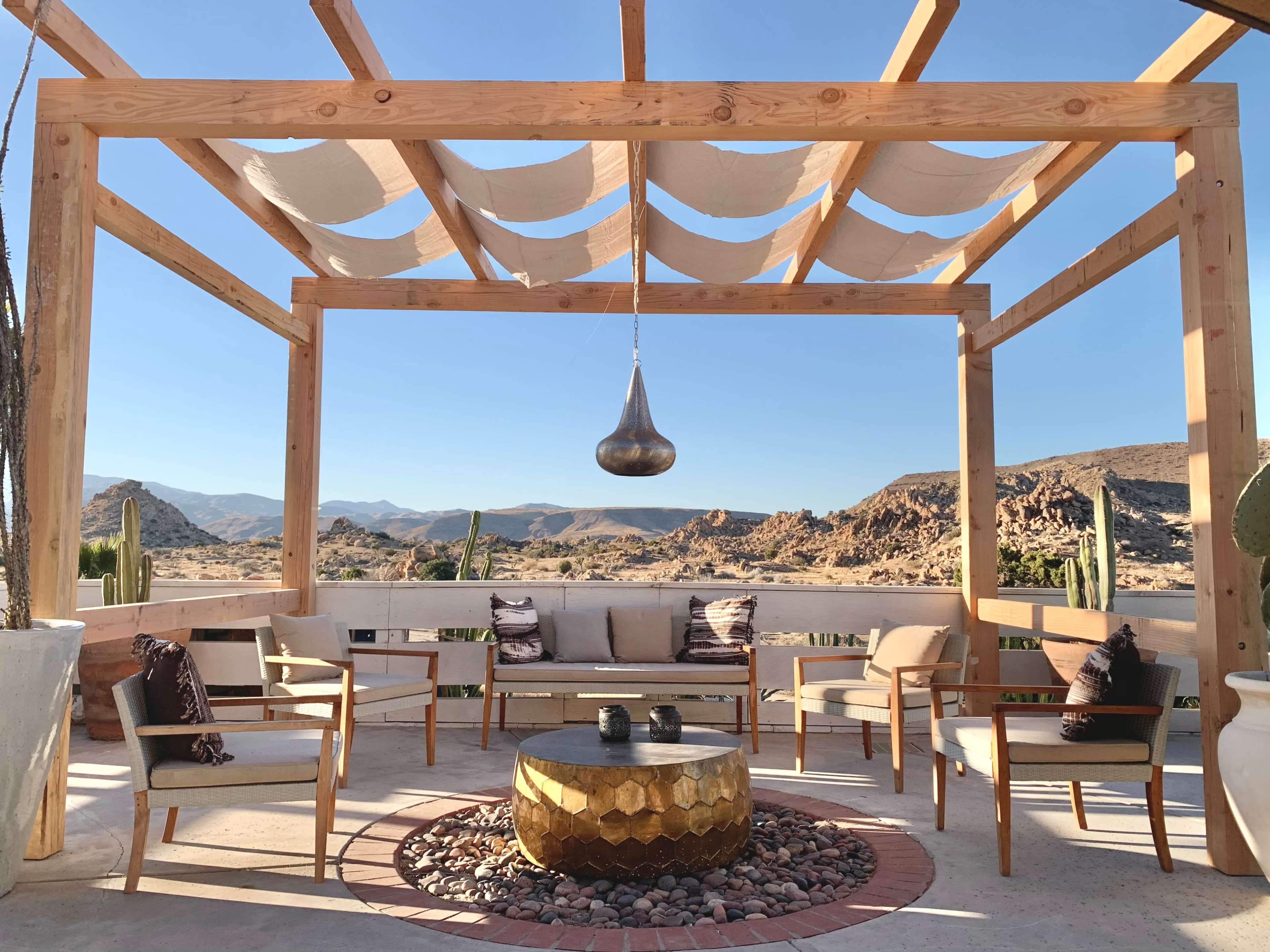 A wooden pergola overlooking a rocky landscape, with a round table surrounded by four chairs and decorative elements like cacti.