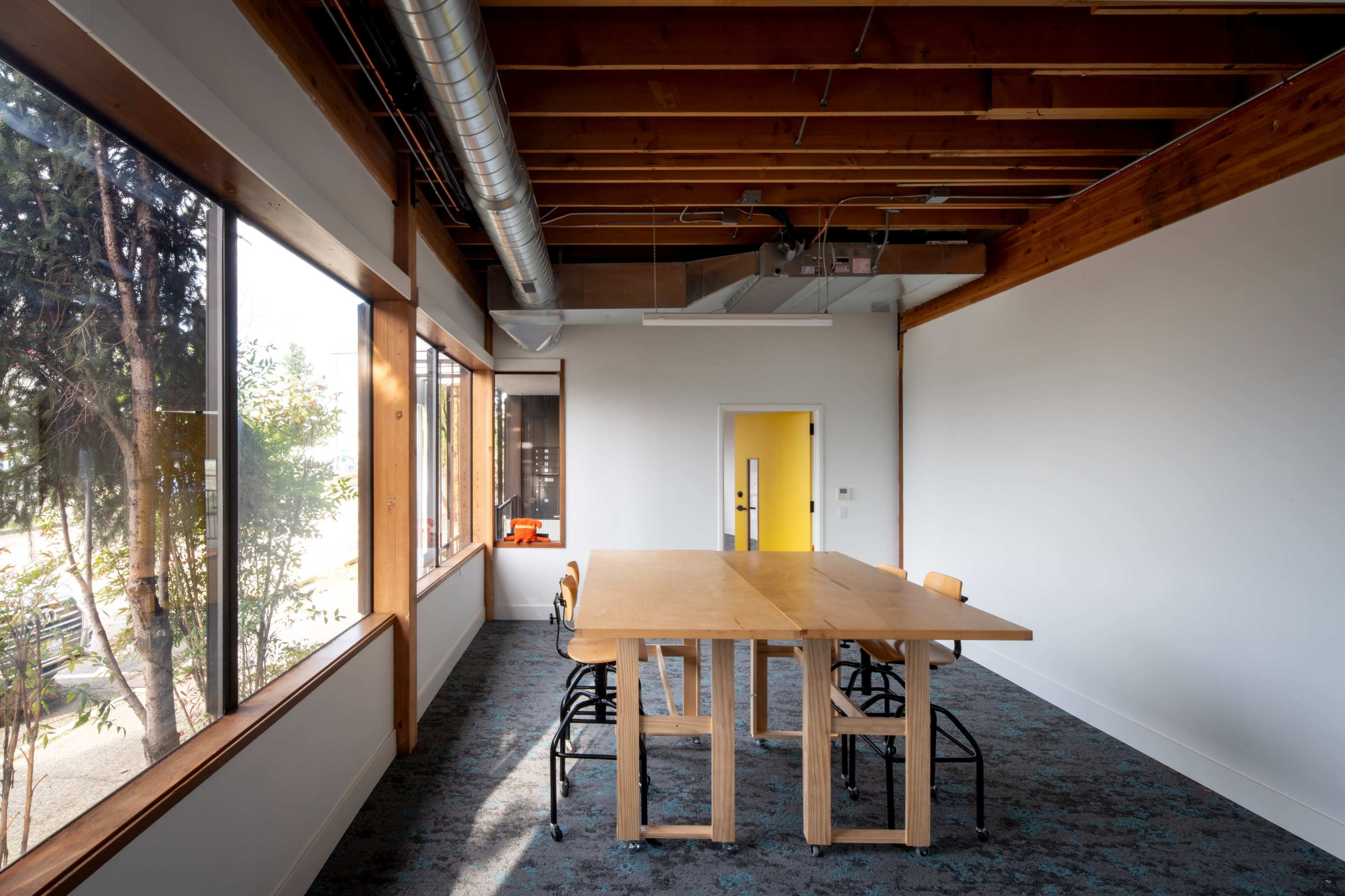 A large wooden table with metal chairs is positioned in a brightly lit conference room featuring large windows and exposed wooden beams.