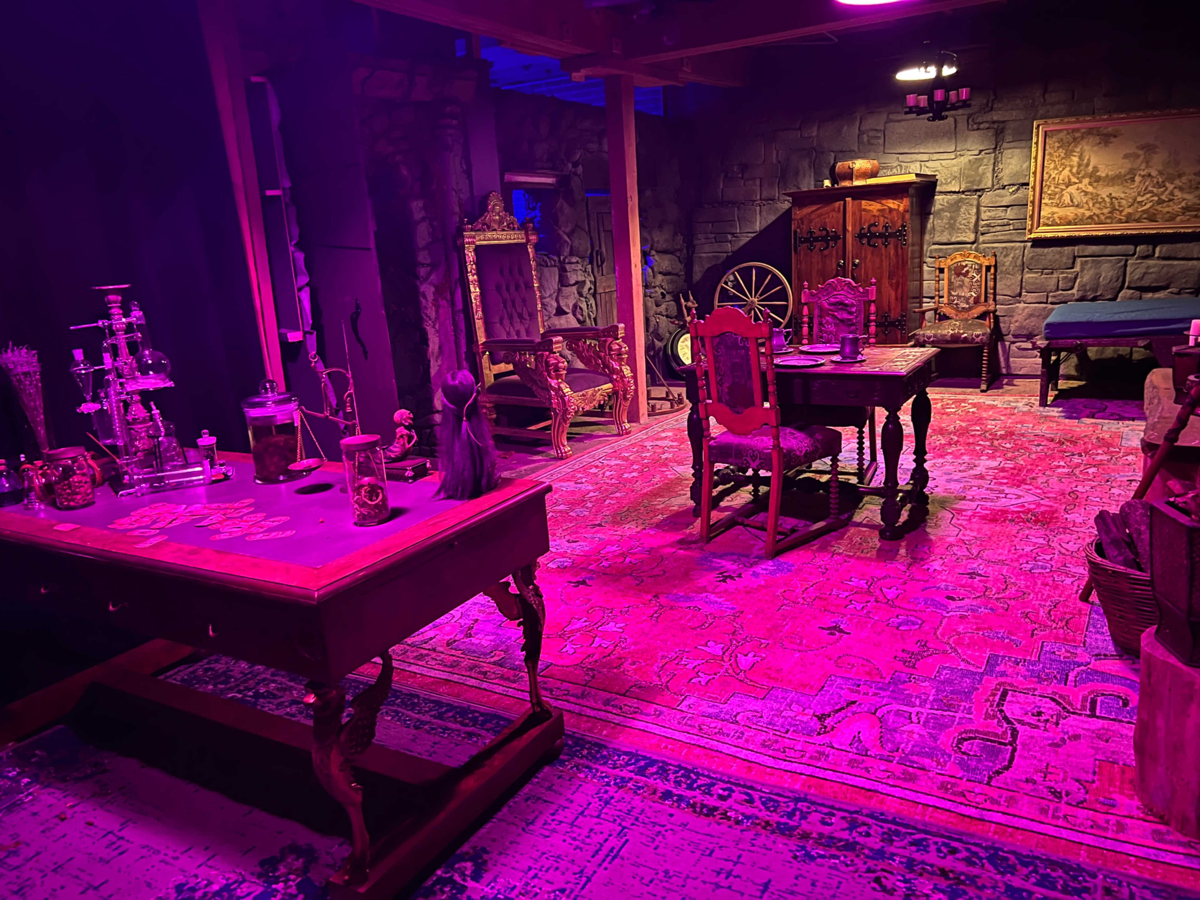 The image shows a dimly lit room with purple lighting, featuring antique furniture including a table, chairs, and a cabinet, set against stone walls and a patterned rug.